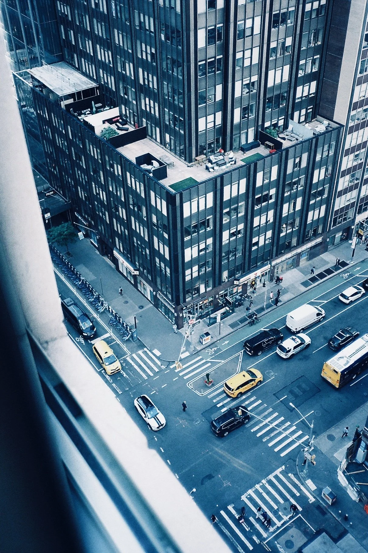 Aerial view of city street with cars, taxis, pedestrians, crosswalks, and a tall modern office building with a rooftop terrace.