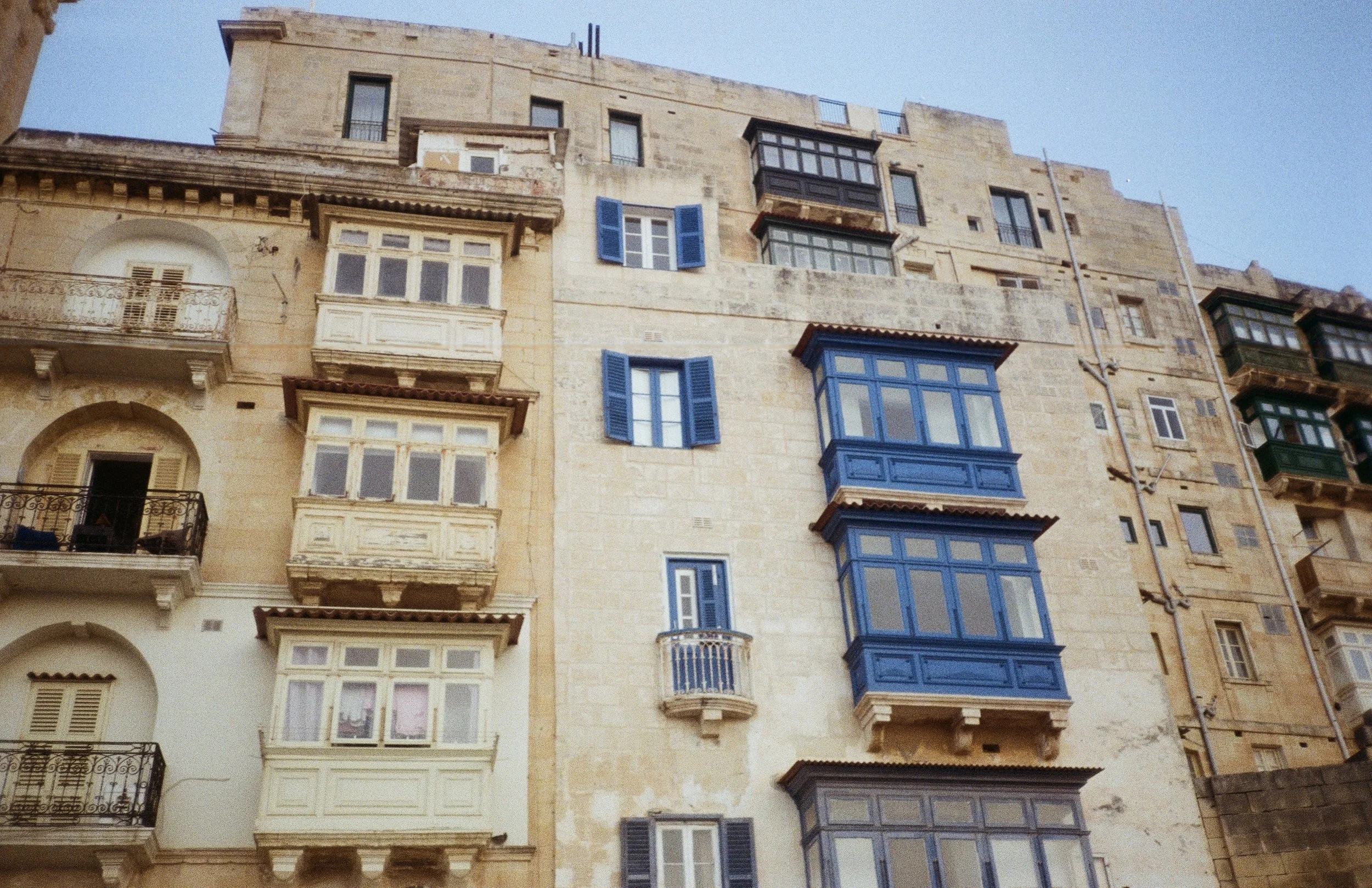 Multi-story stone building with various enclosed balconies and windows, some with blue shutters or framing, under a clear blue sky.