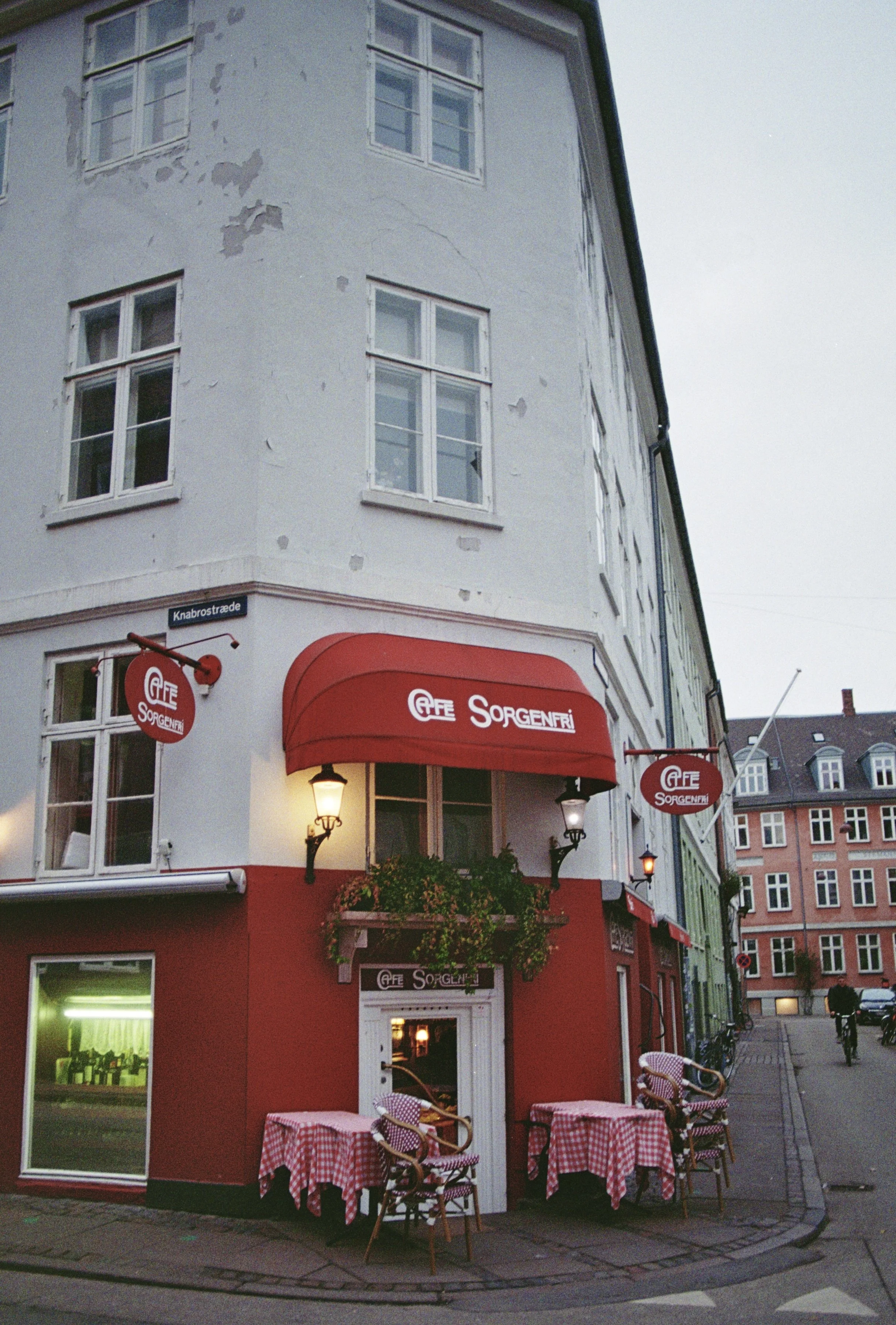 Corner of a building with a red awning and outdoor seating with checkered tablecloths, signs reading 'Cafe Sorgenfri,' with bicycles and pedestrian nearby on a city street.