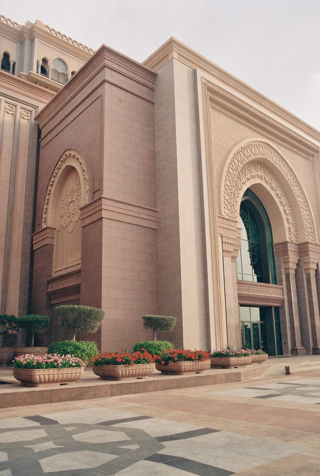 Exterior of a large, ornate building with pink and beige tones, arched windows, and decorative details; potted plants with flowers and trimmed bushes are in front.