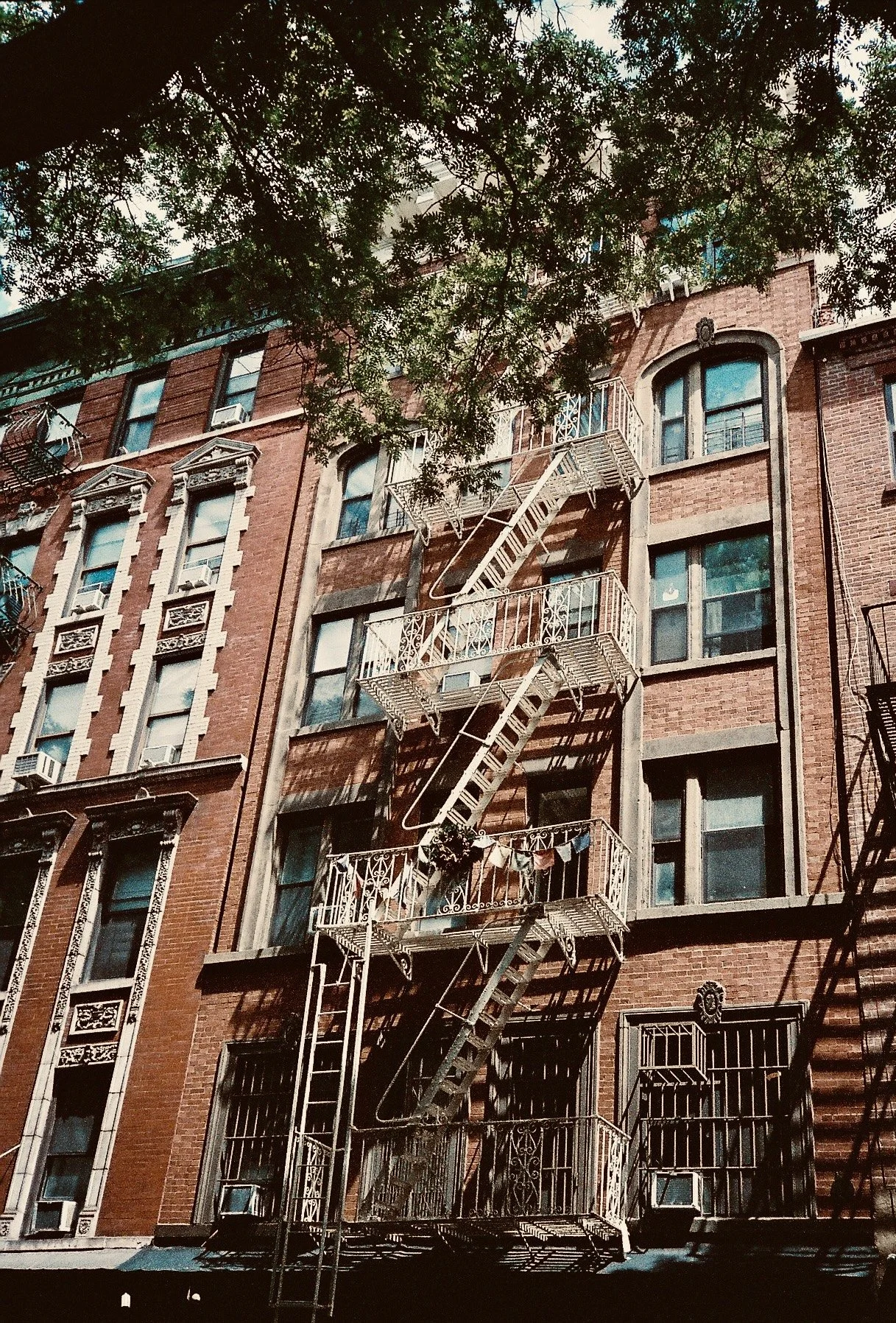 A brick apartment building with fire escape stairs and laundry hanging on one of the balconies, with trees in the foreground.