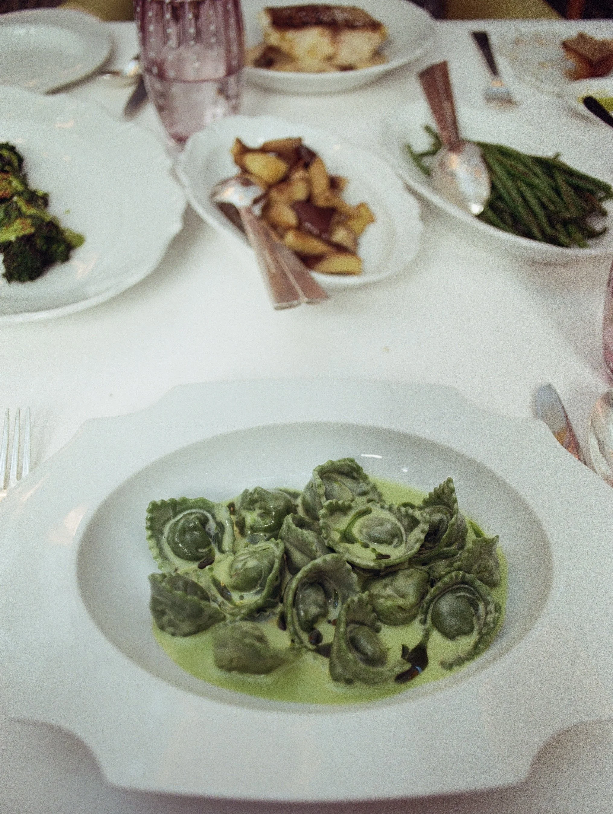 Plate of cooked orecchiette pasta with green sauce on a white table, surrounded by side dishes including roasted potatoes, green beans, and broccoli, with glasses of water, cutlery, and napkins.