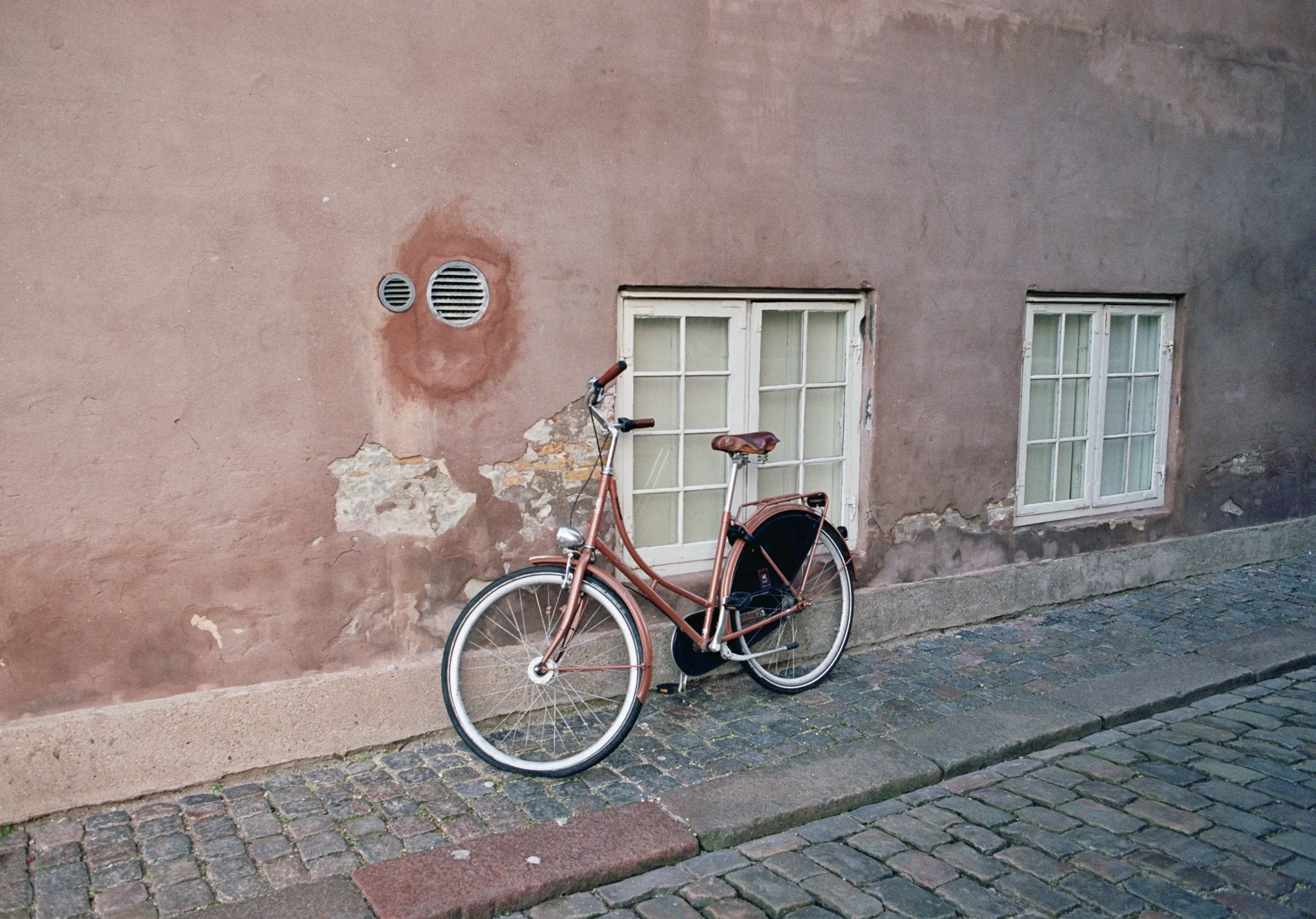 A vintage bicycle with a brown leather seat leaning against a weathered pink wall with peeling paint, two small windows, and two round air vents, on a cobblestone street.