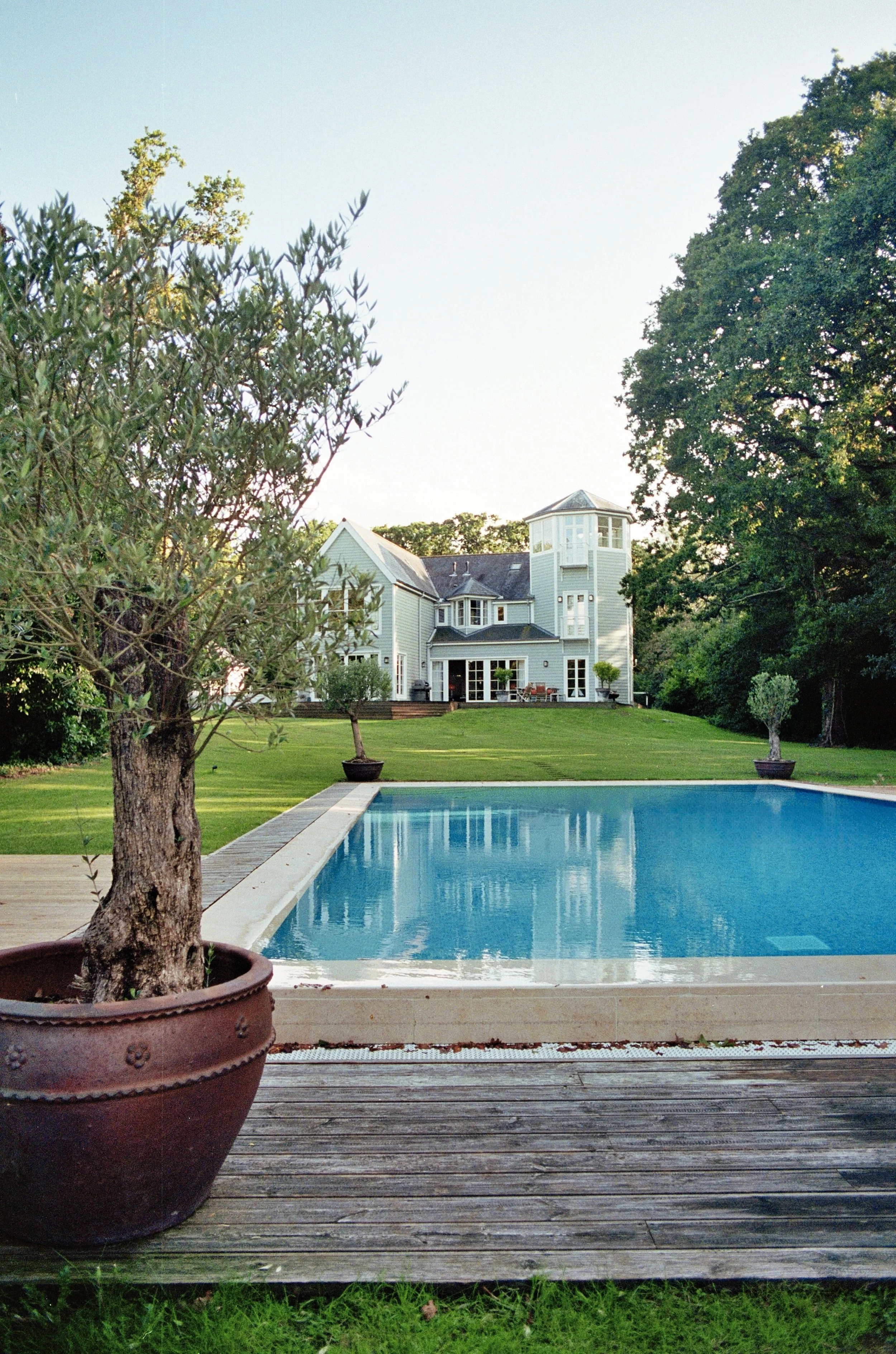 Backyard with swimming pool, potted trees, and large white house with turret and porch.