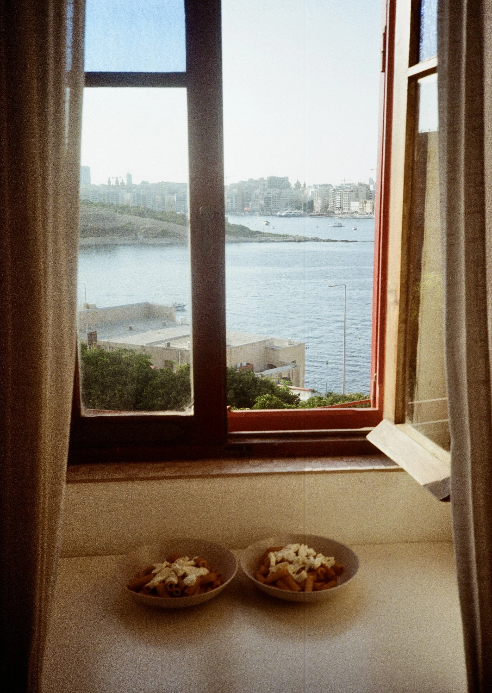 Two bowls of pasta placed on a windowsill with a view of a river, boats, and city buildings through an open window with curtains.