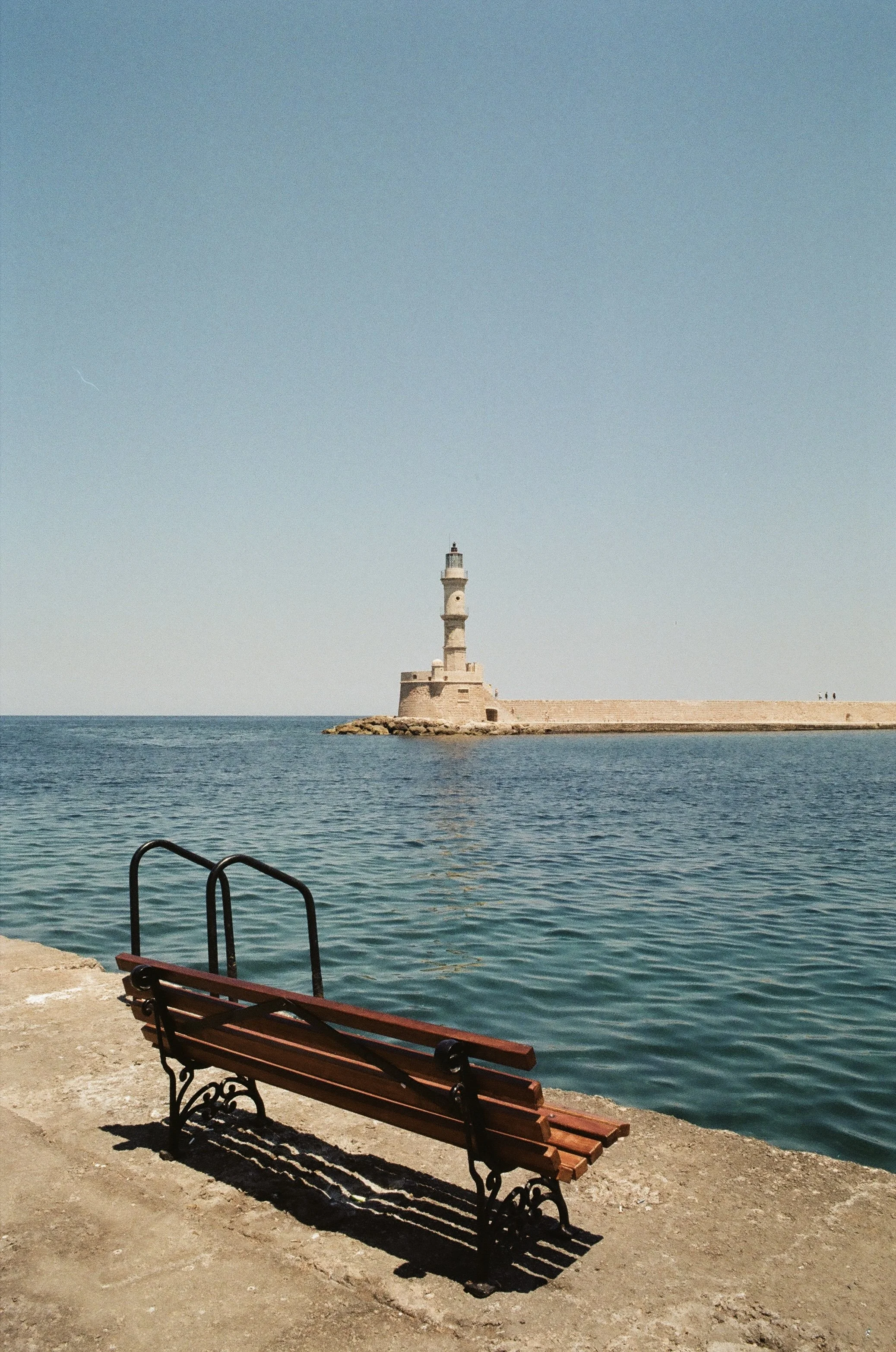 A seaside scene with a wooden bench in the foreground, clear blue water, and a lighthouse on a breakwater in the distance under a bright blue sky.