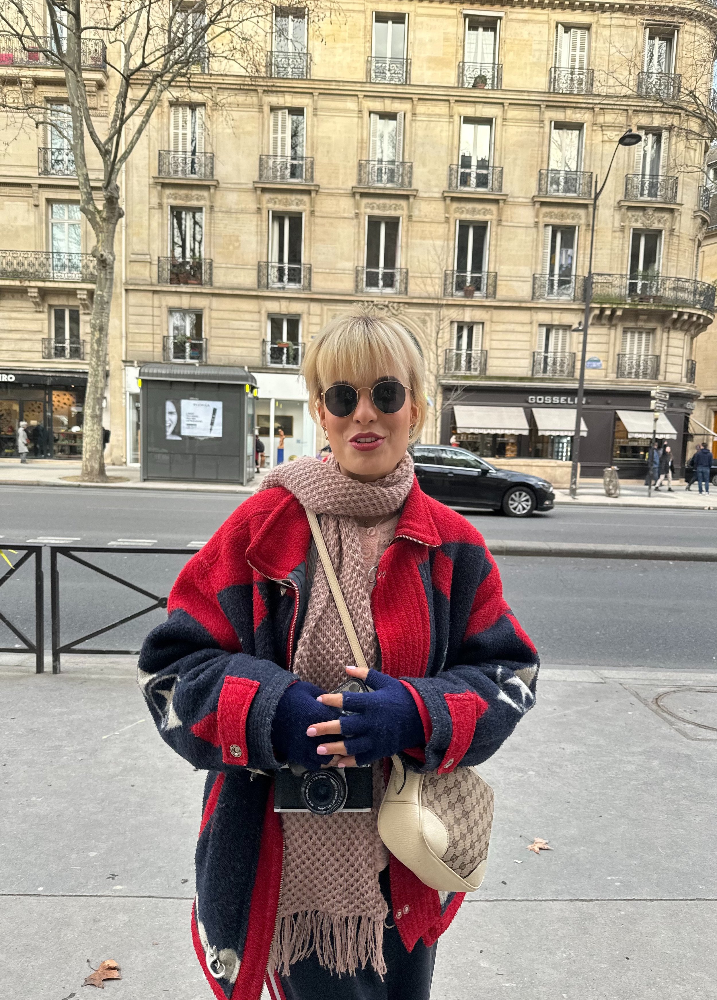 A young woman with blond hair and sunglasses standing on a city sidewalk, wearing a red, navy, and cream patterned coat, a beige scarf, and gloves. She has a small crossbody bag and a camera hanging from her neck. Behind her are typical Parisian buildings with balconies, and people walking along the street.