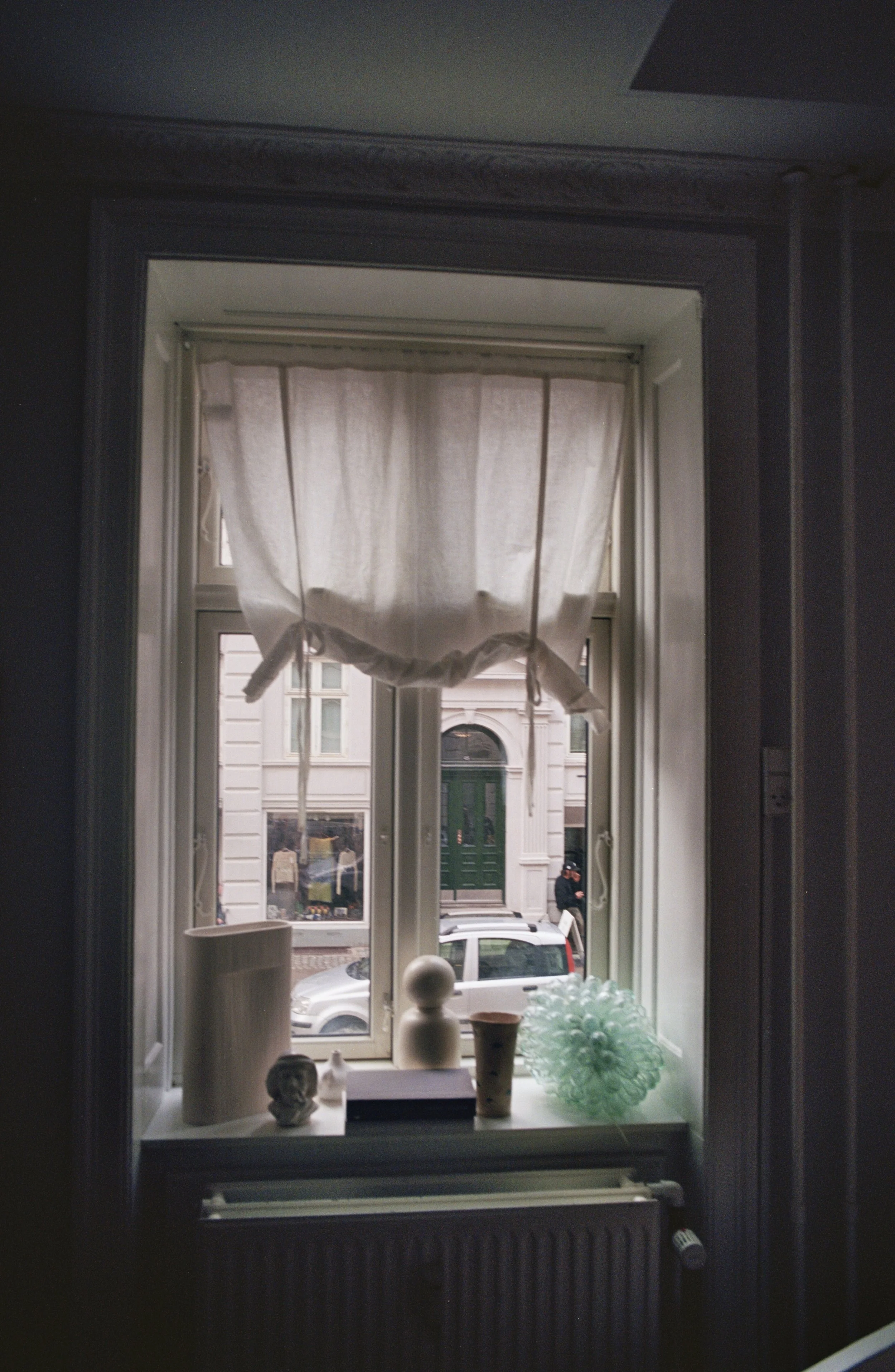 View through a window showing a street scene with a parked silver car and pedestrians outside, decorated with white curtains, home decor items on the windowsill, including a green paper star, and a building with a green door across the street.