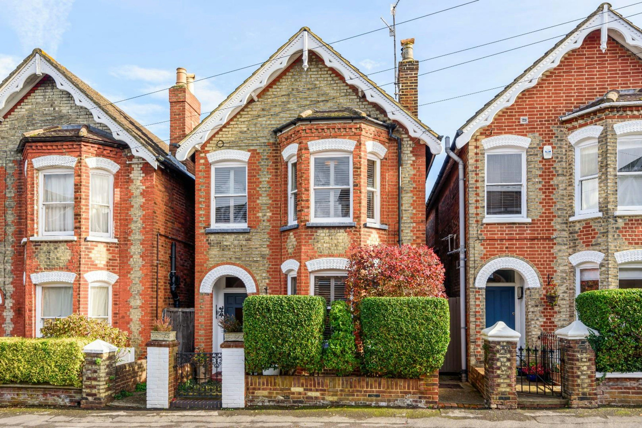 Row of brick and stone Victorian-style houses with front gardens and hedges, under a blue sky.