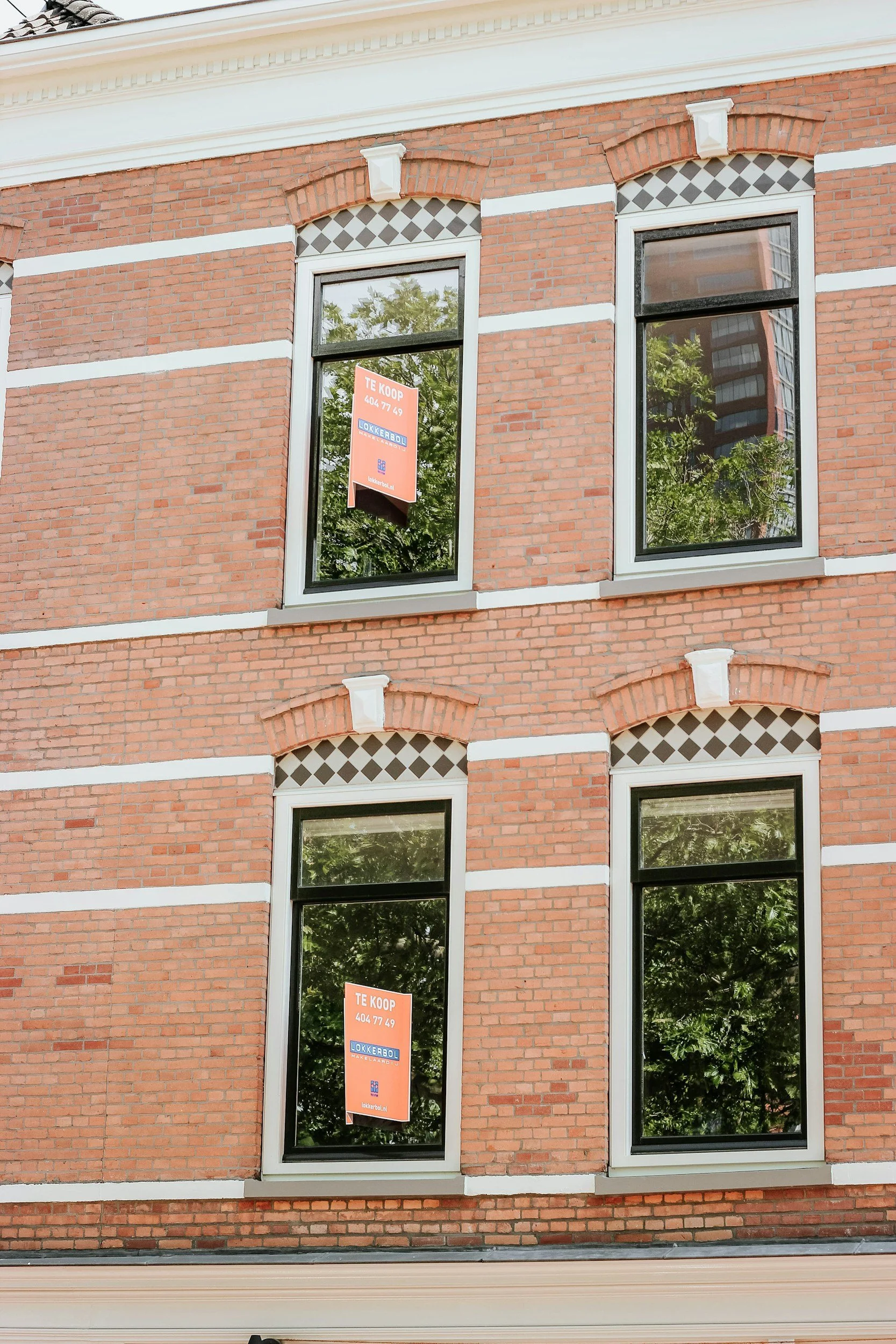 The image shows the exterior of a brick building with four large windows, each with white trim and decorative brick arches above. Two of the windows have orange "For Sale" signs in them that say "TE KOOP" with a contact number and website.
