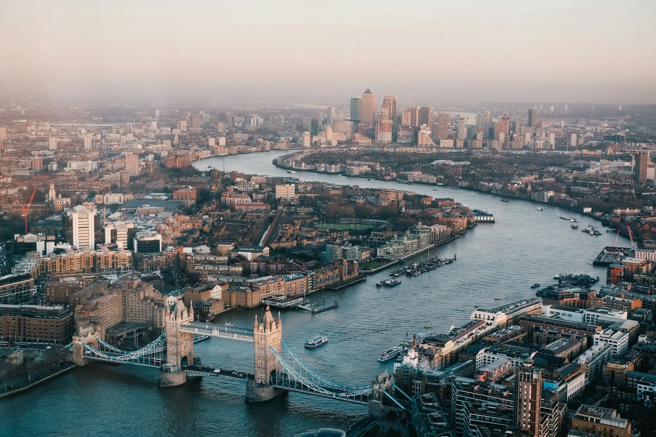 An aerial view of London cityscape featuring Tower Bridge over the River Thames with high-rise buildings and the skyline in the background during sunset.