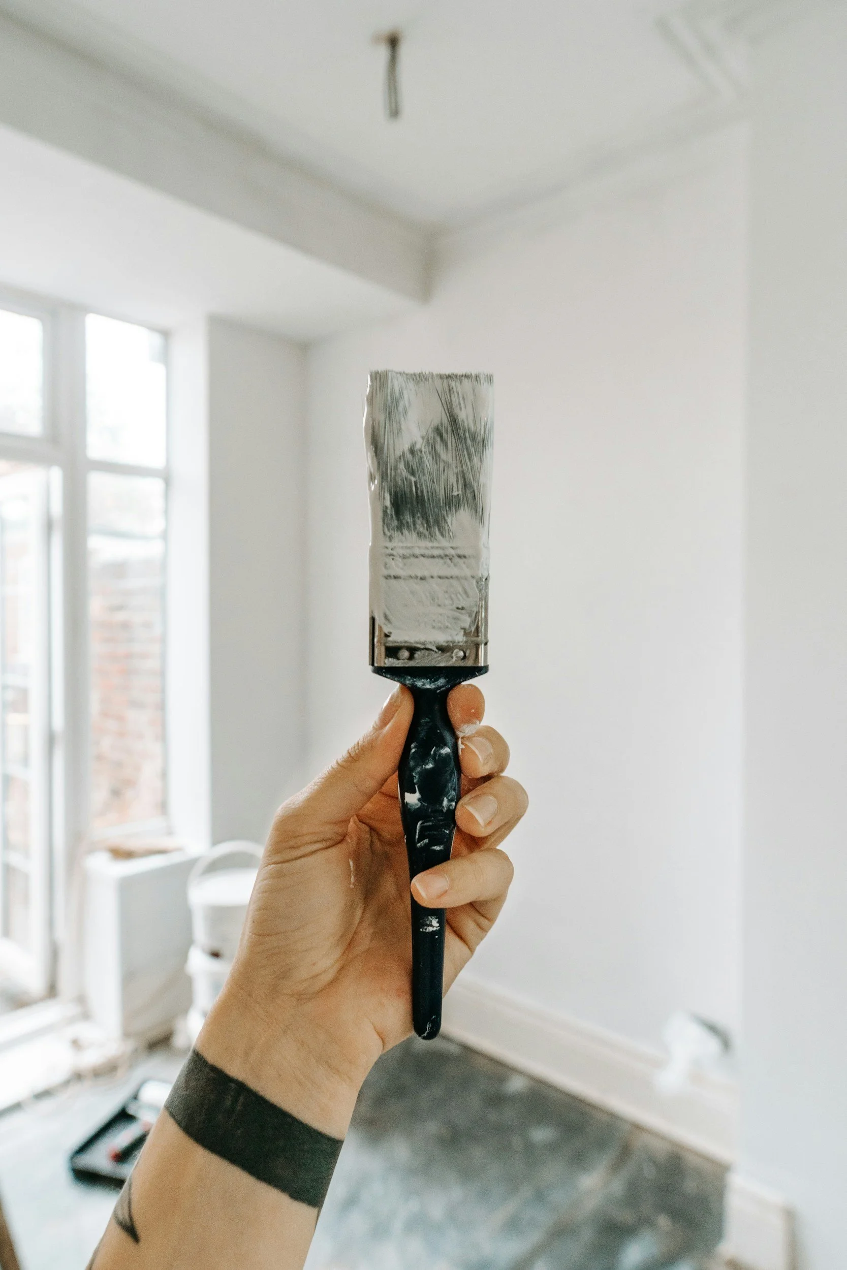 A hand holding a paintbrush with wet silver paint inside a room with white walls and a window.