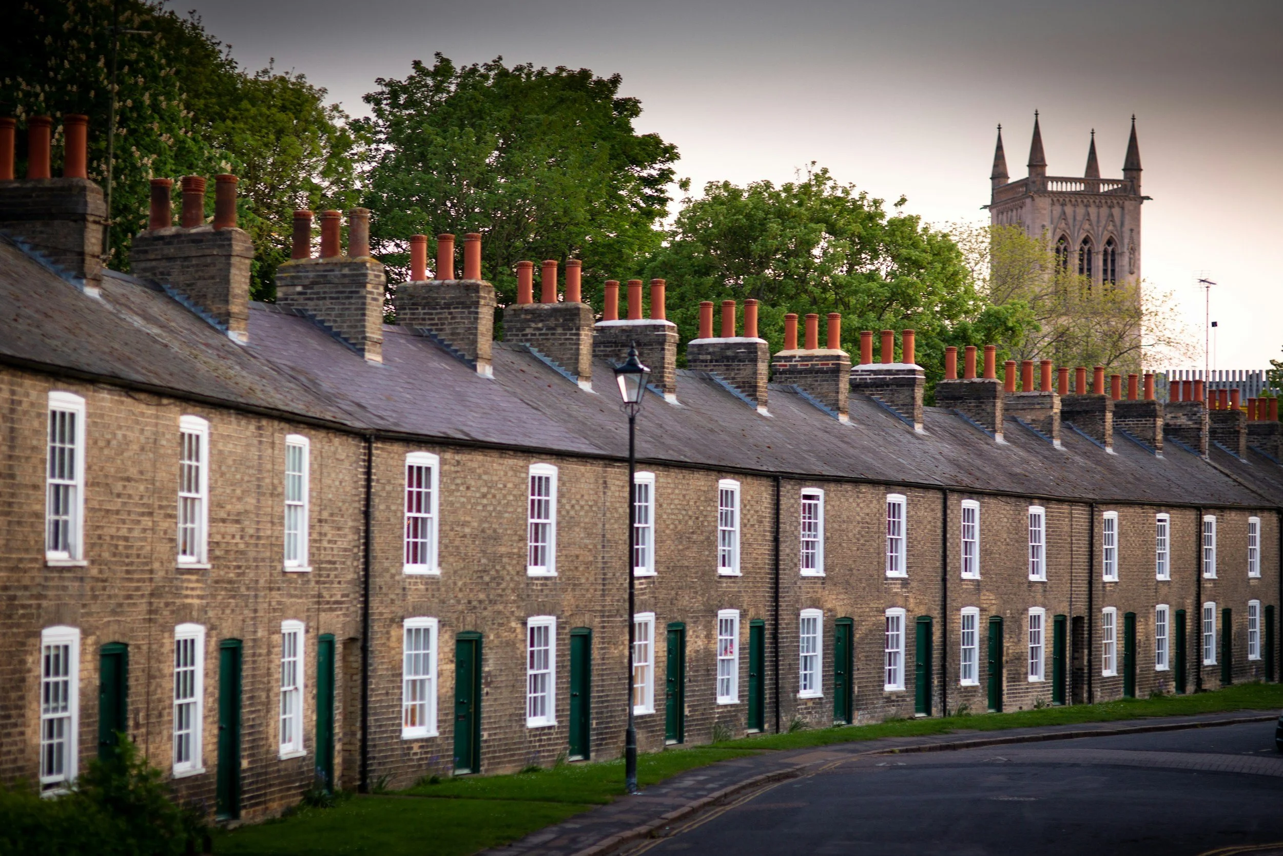 Row of traditional brick houses with green doors and white-framed windows, chimney stacks, street lamp, and church tower in the background.