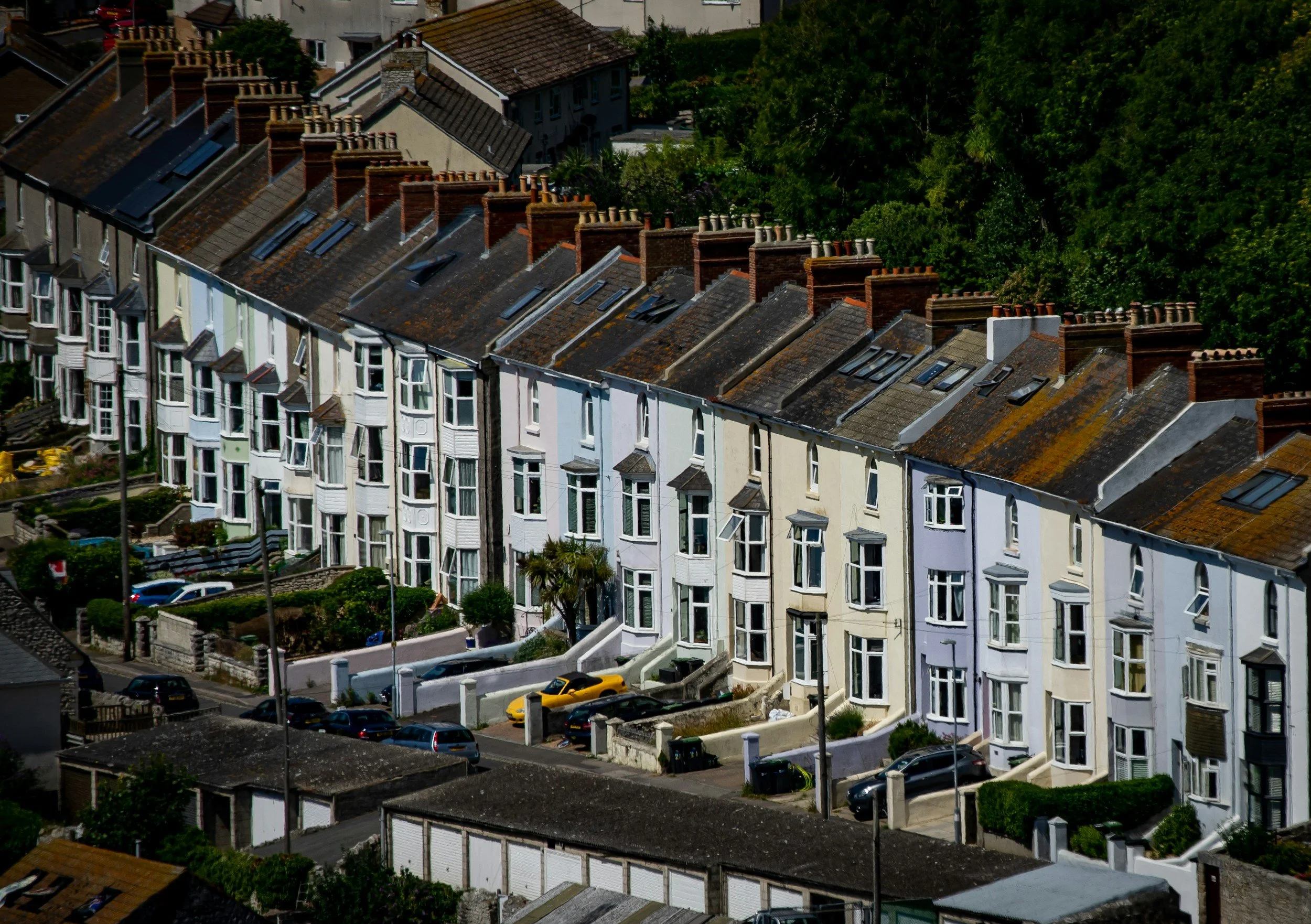 A row of pastel-colored Victorian-style terraced houses with bay windows, parked cars, and chimneys on the rooftops, with green trees in the background.