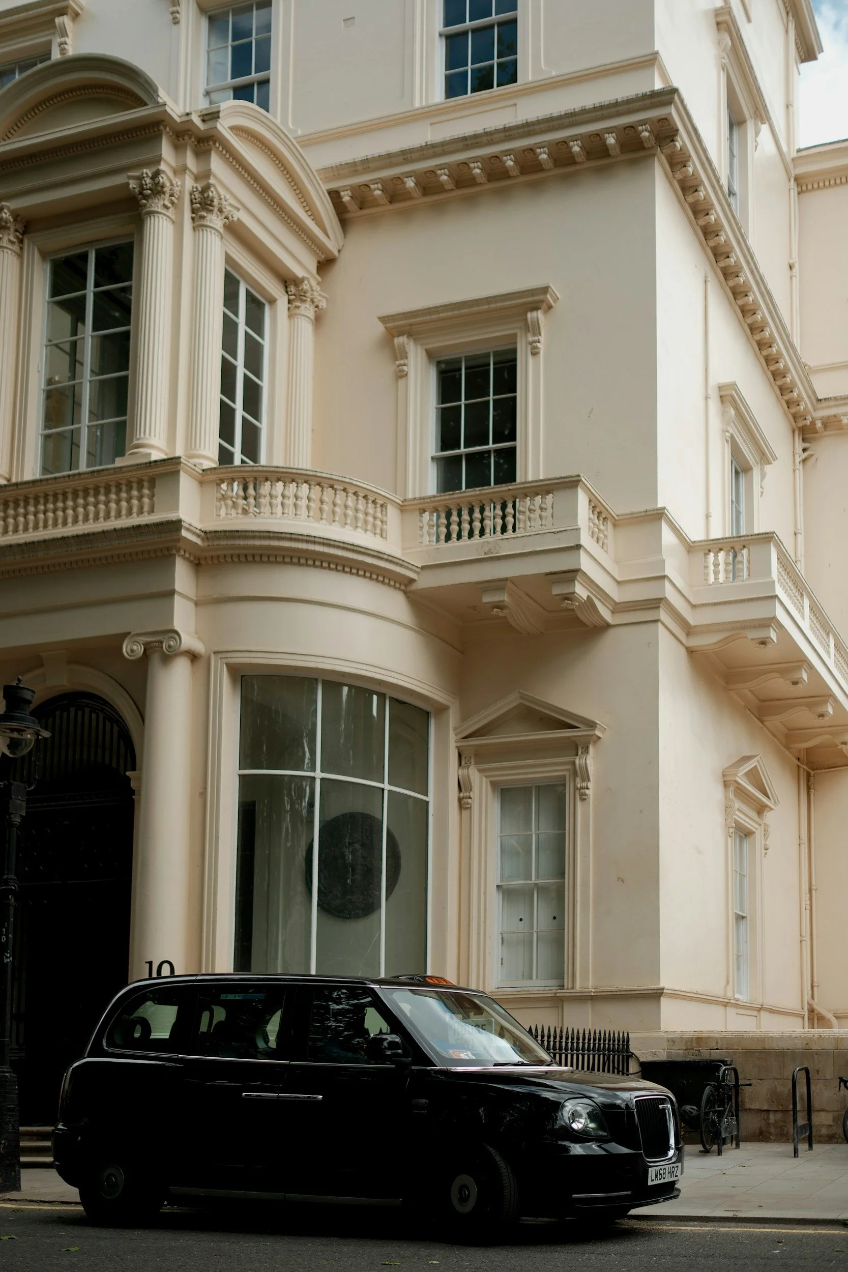 A black London taxi cab parked in front of an elegant, cream-colored building with ornate architectural details and large windows.