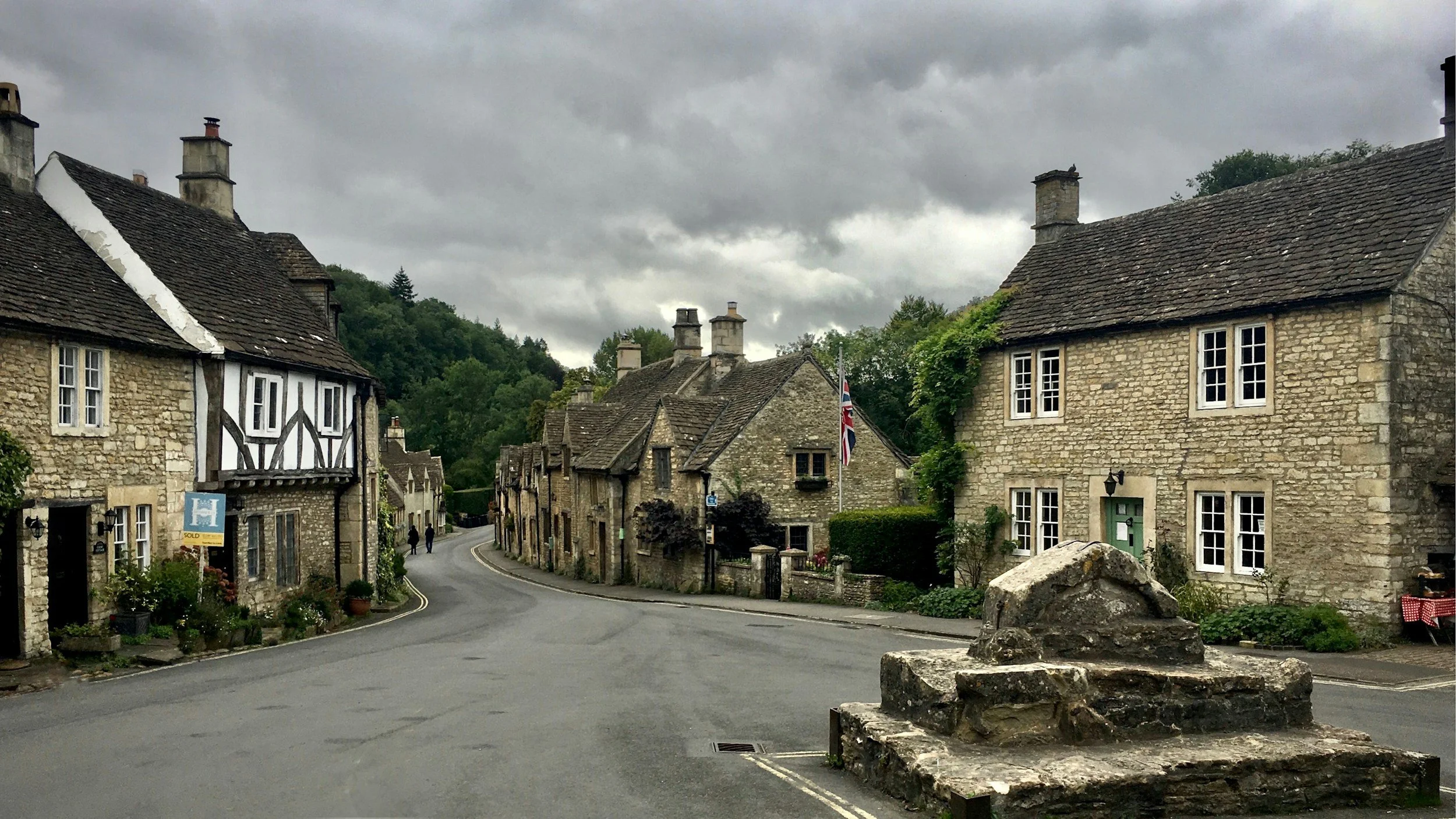 A quiet, curving street in a small British village, with historic stone cottages, a cloudy sky, and greenery in the background.