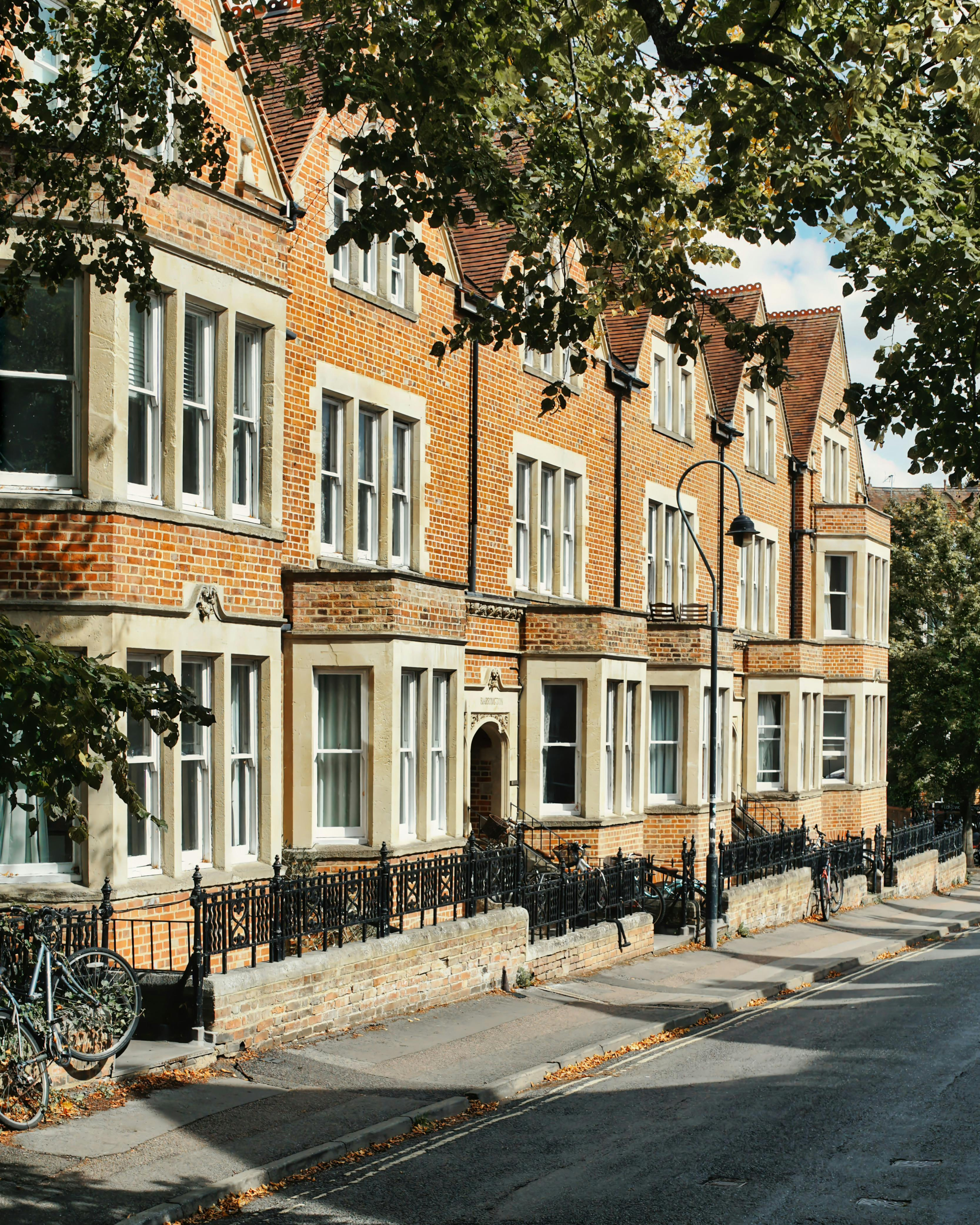 A row of pastel-colored Victorian-style terraced houses with bay windows, parked cars, and chimneys on the rooftops, with green trees in the background.
