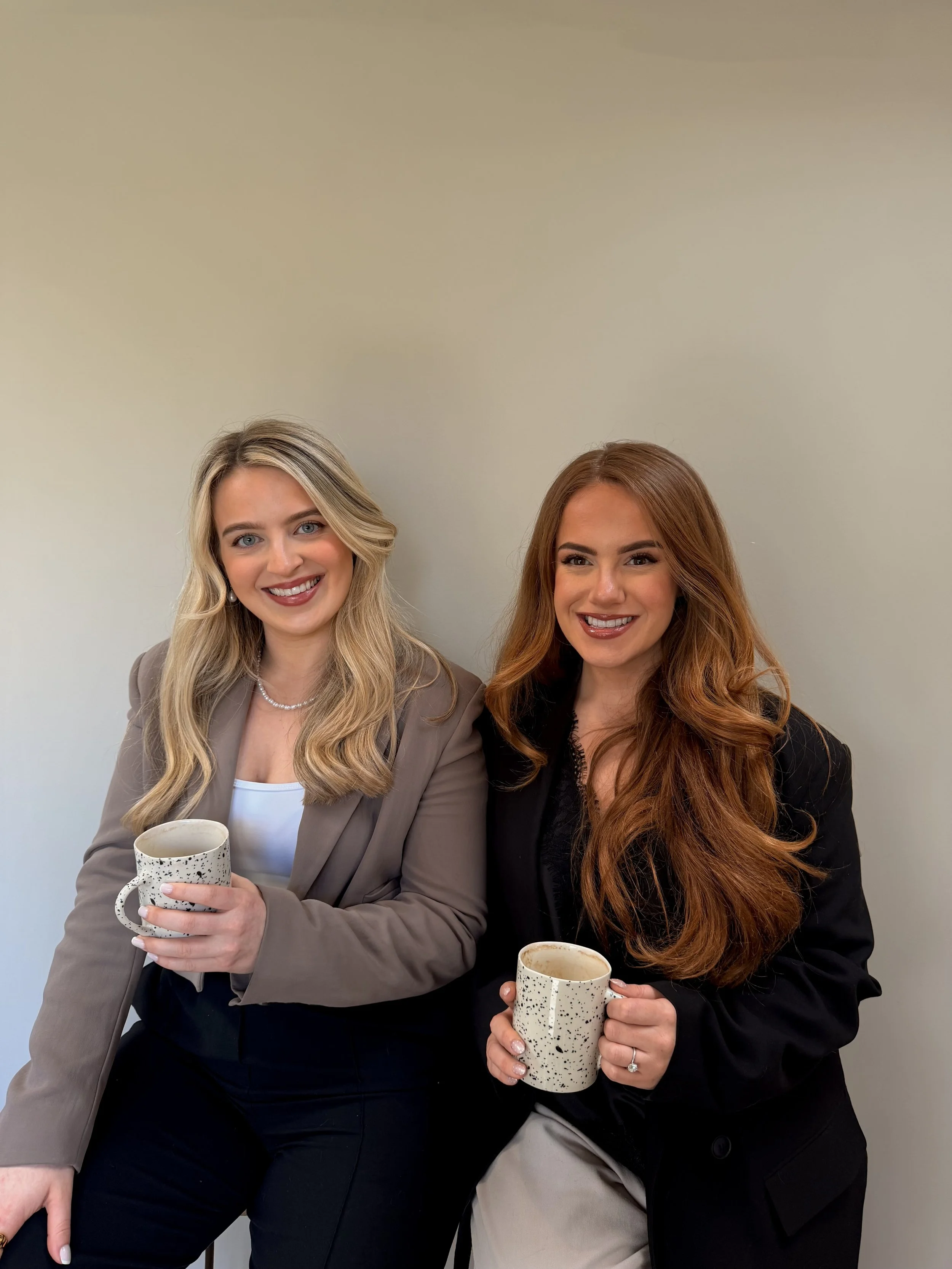 Two women smiling and holding coffee mugs, standing in front of a plain, light-colored wall.