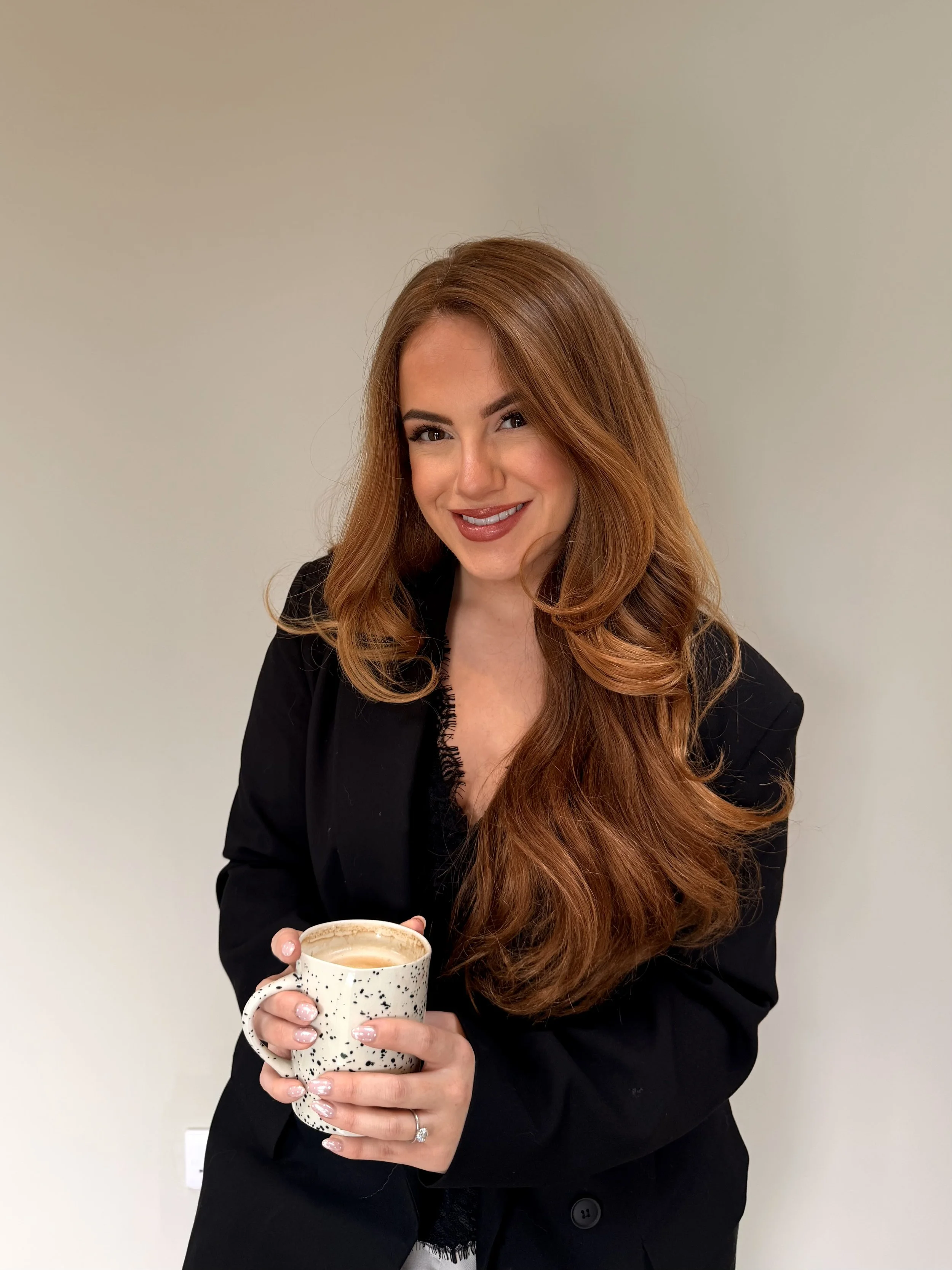 A woman with long red hair, smiling, holding a mug of coffee or tea, wearing a black blazer against a plain light-colored background.