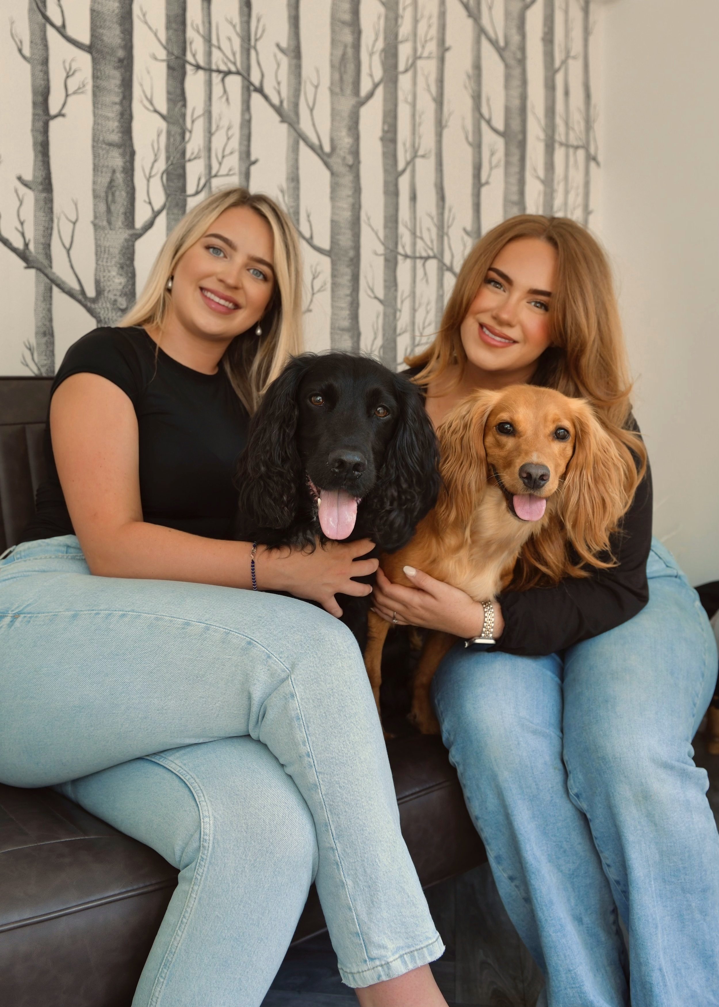 Two women sitting on a couch holding two dogs in front of a wall with a tree pattern
