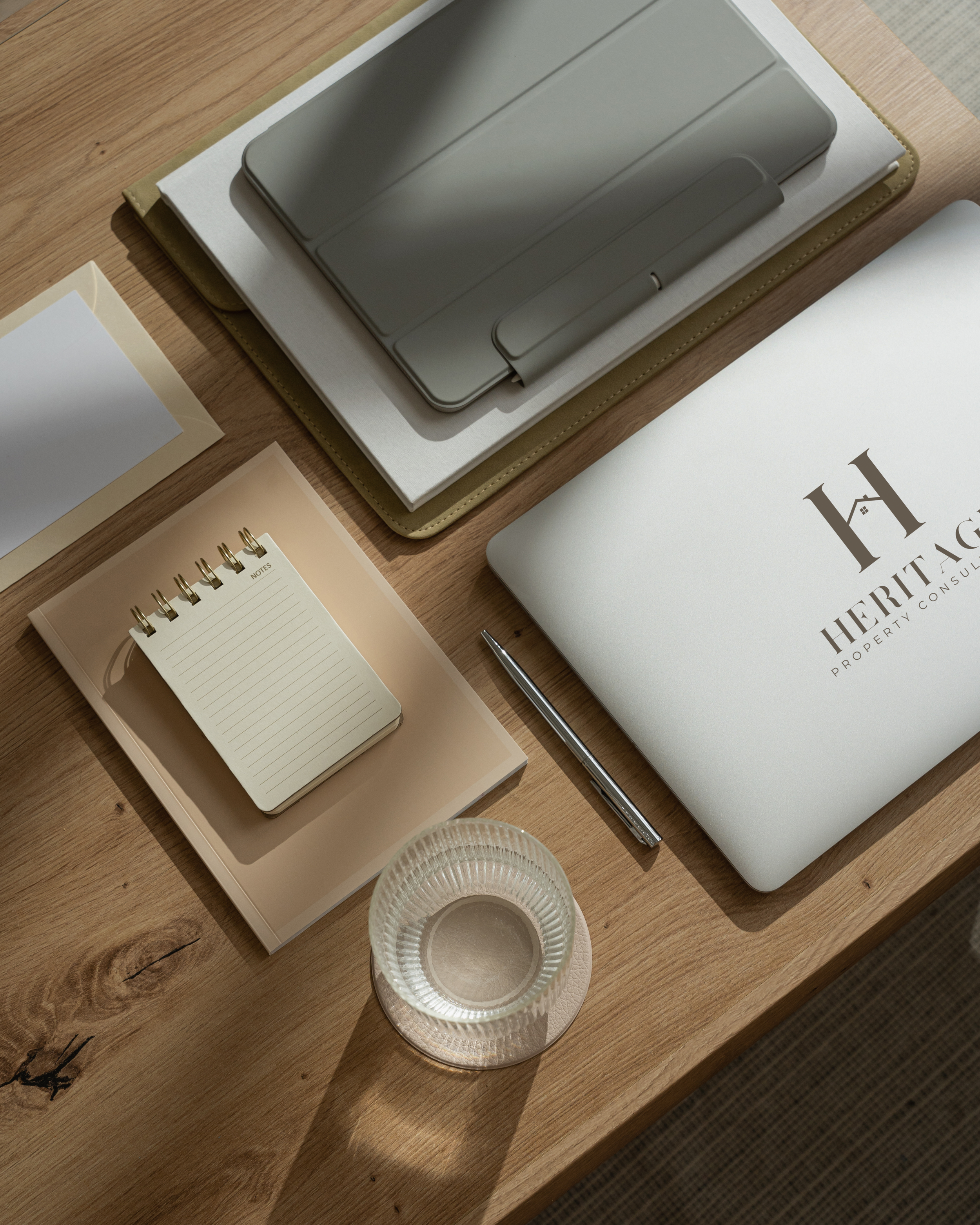 Two people sitting at a round white table using laptops and a smartphone, with a small potted plant, a takeaway coffee cup, a notebook, and a tablet on the table. Sunlight coming through a window.