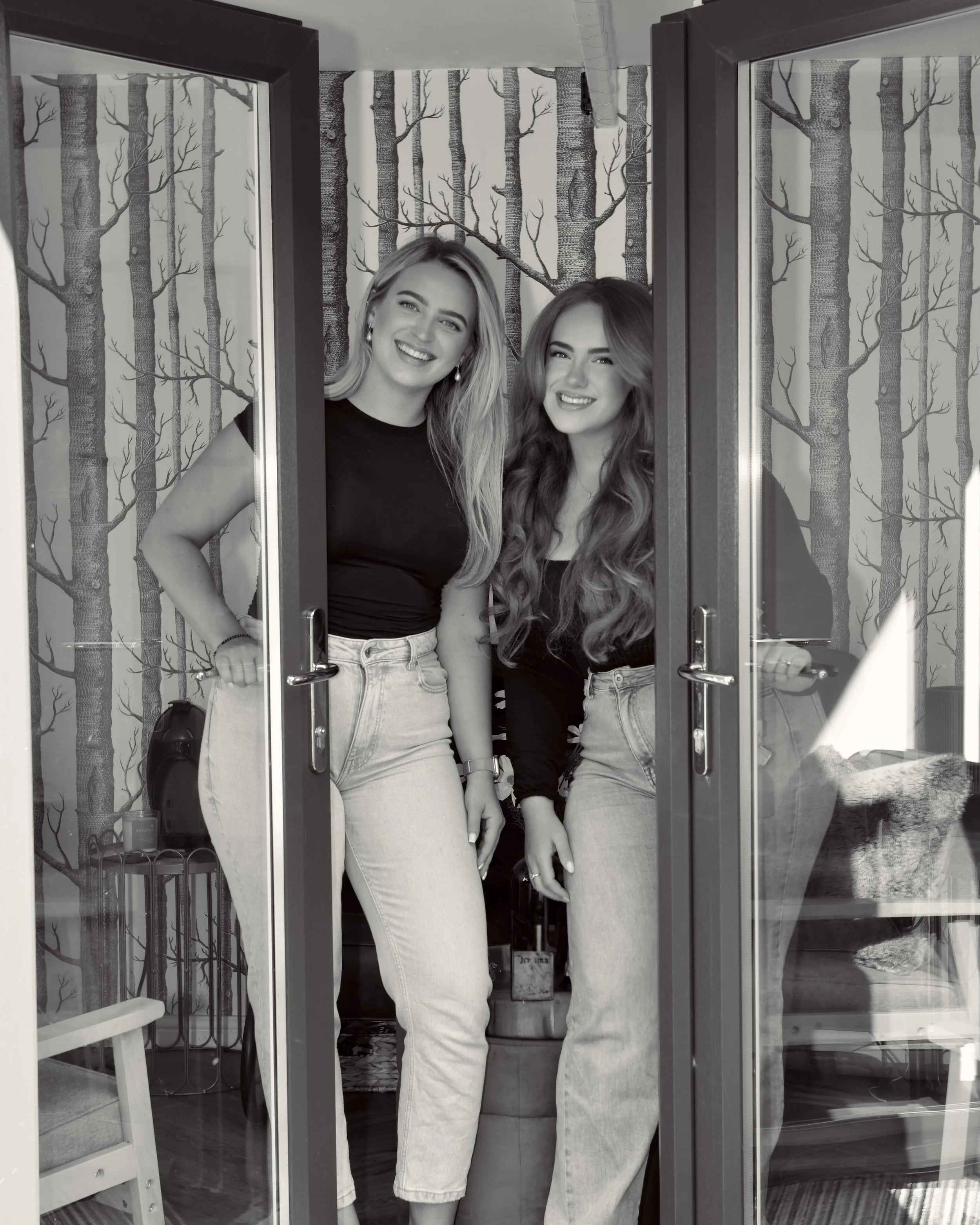 Two young women with long hair smiling and standing inside a room, seen through glass doors, with a forest-themed wallpaper in the background.
