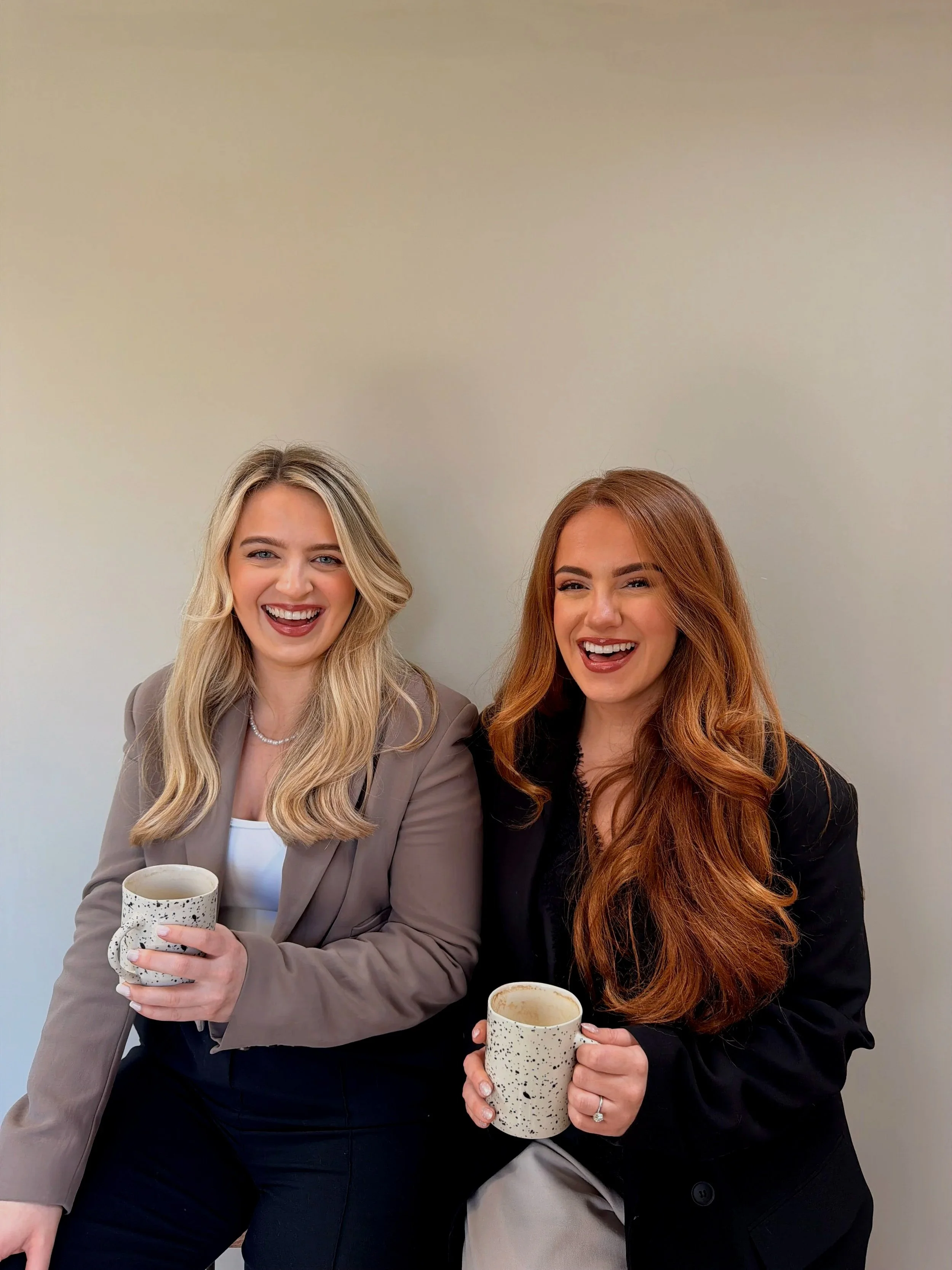 Two women sitting together, smiling and holding speckled ceramic mugs, against a plain beige wall.