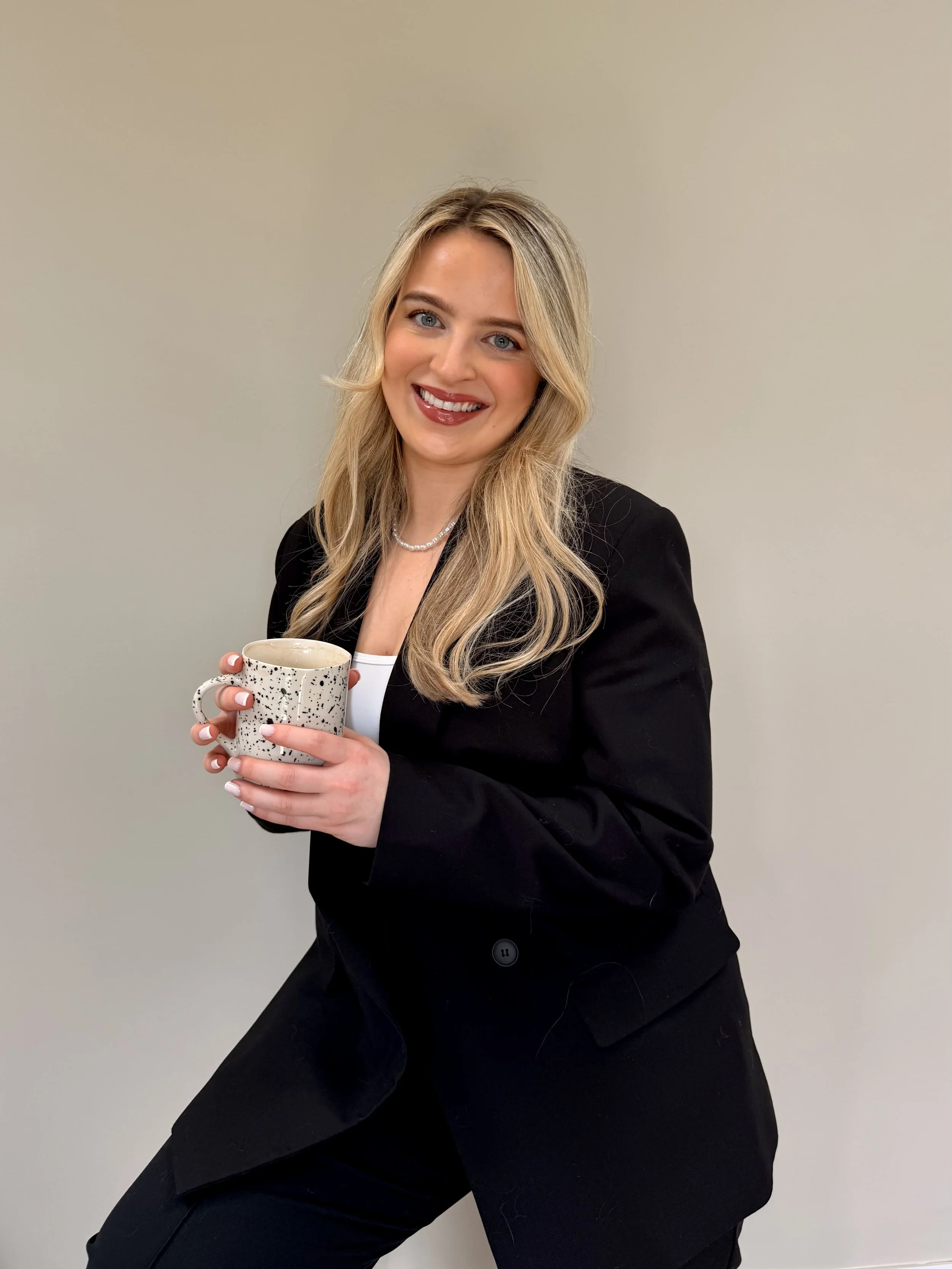 A woman with long blonde hair smiling and holding a mug, standing against a plain light gray background.