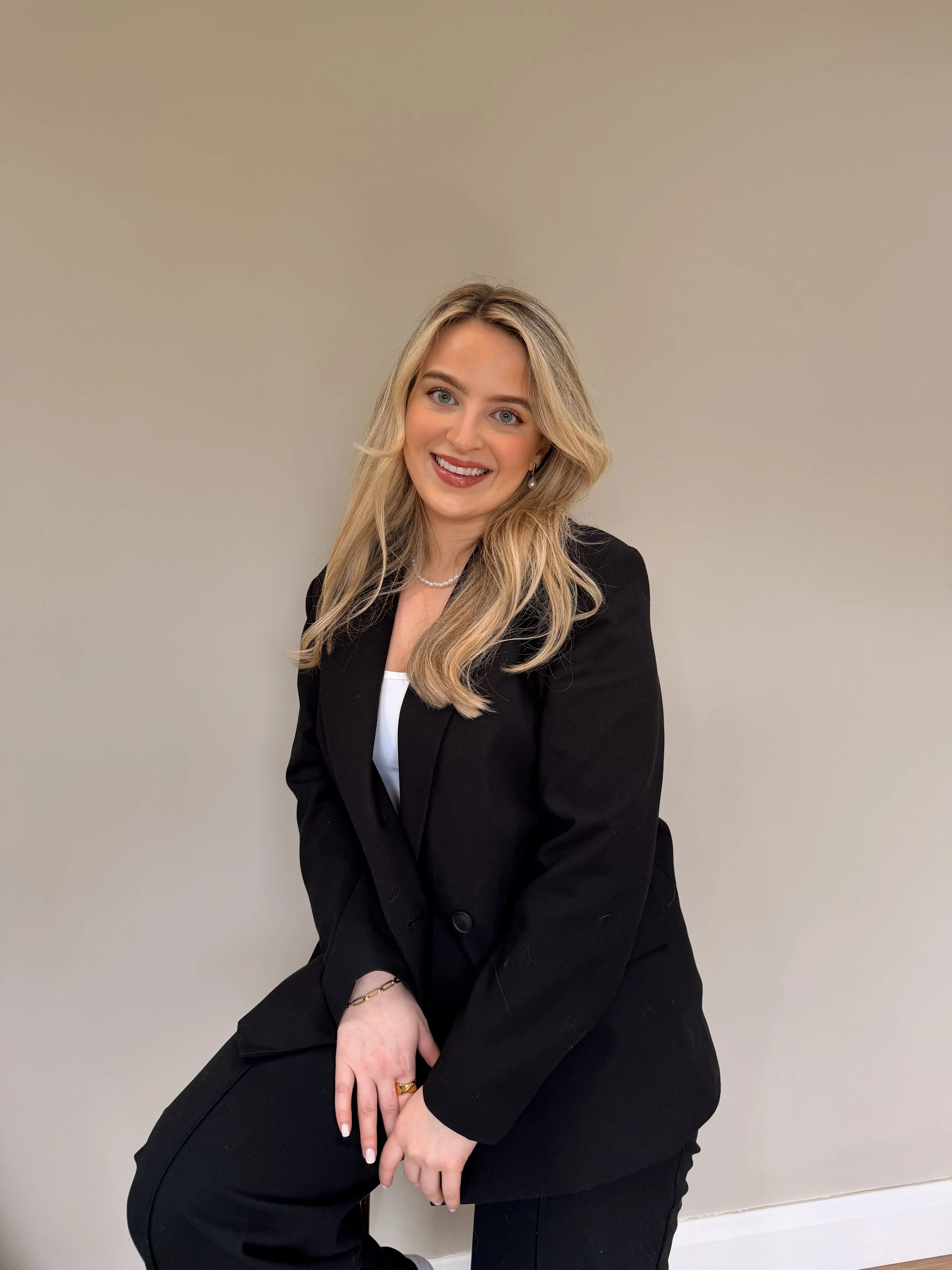A young woman with blonde hair and blue eyes sitting against a plain, light-colored wall, smiling at the camera. She is dressed in a black blazer and white top, accessorized with a pearl necklace, earrings, and rings.