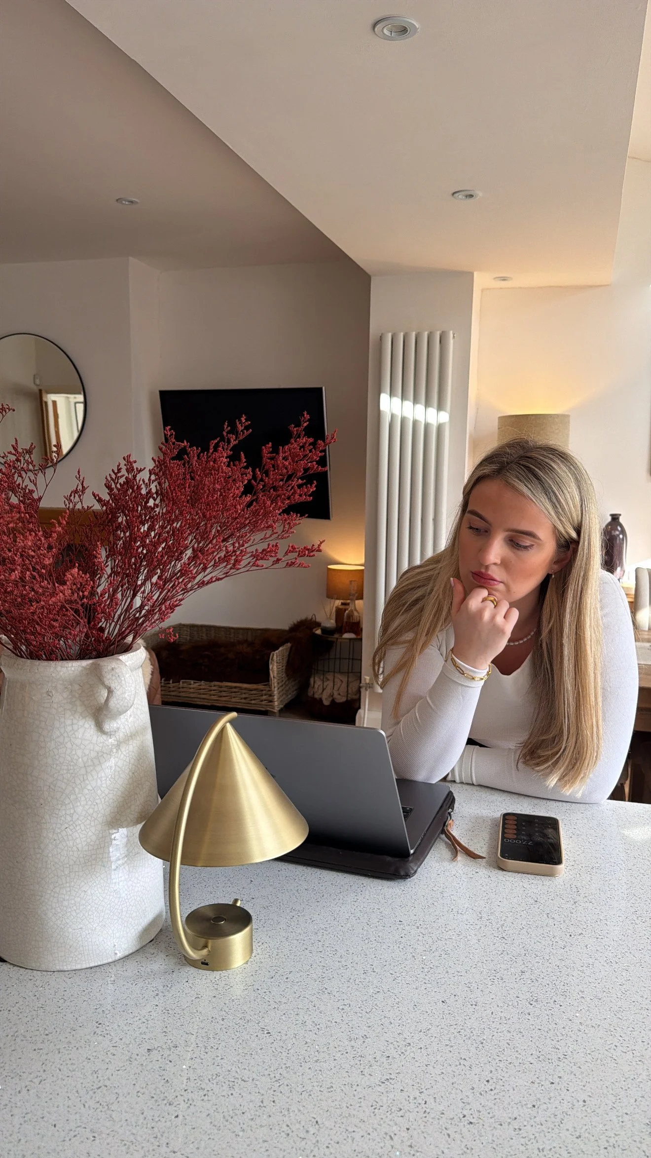 A woman sitting at a kitchen counter with a laptop, phone, and decorative vase with pink flowers, in a cozy living room setting.