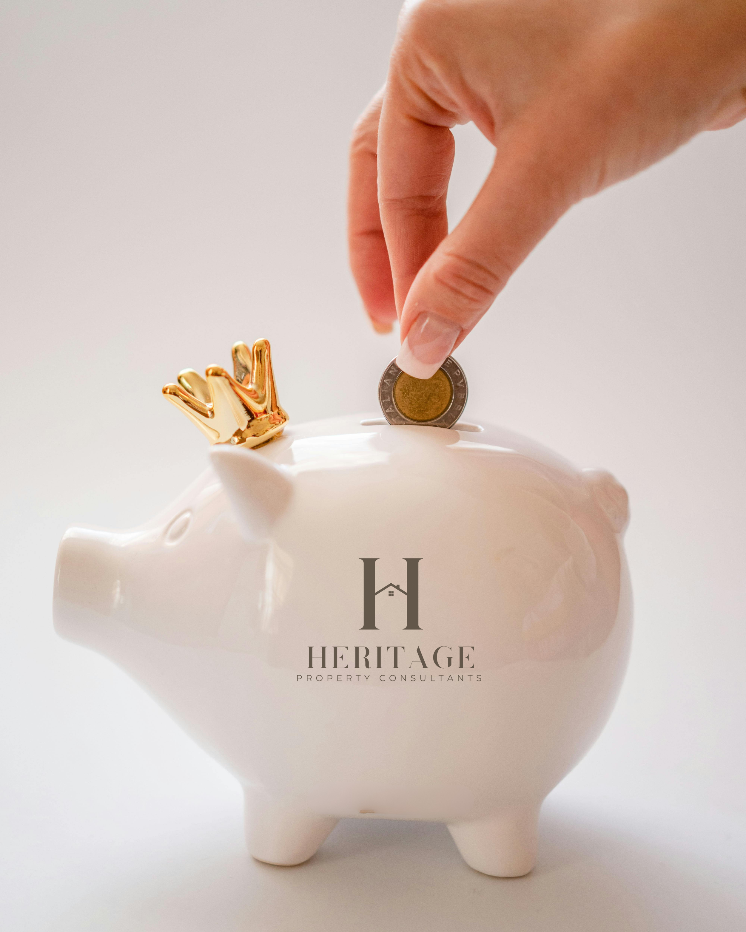 Person inserting a coin into a white piggy bank with a gold crown on its back, featuring the logo 'Heritage Property Consultants'.