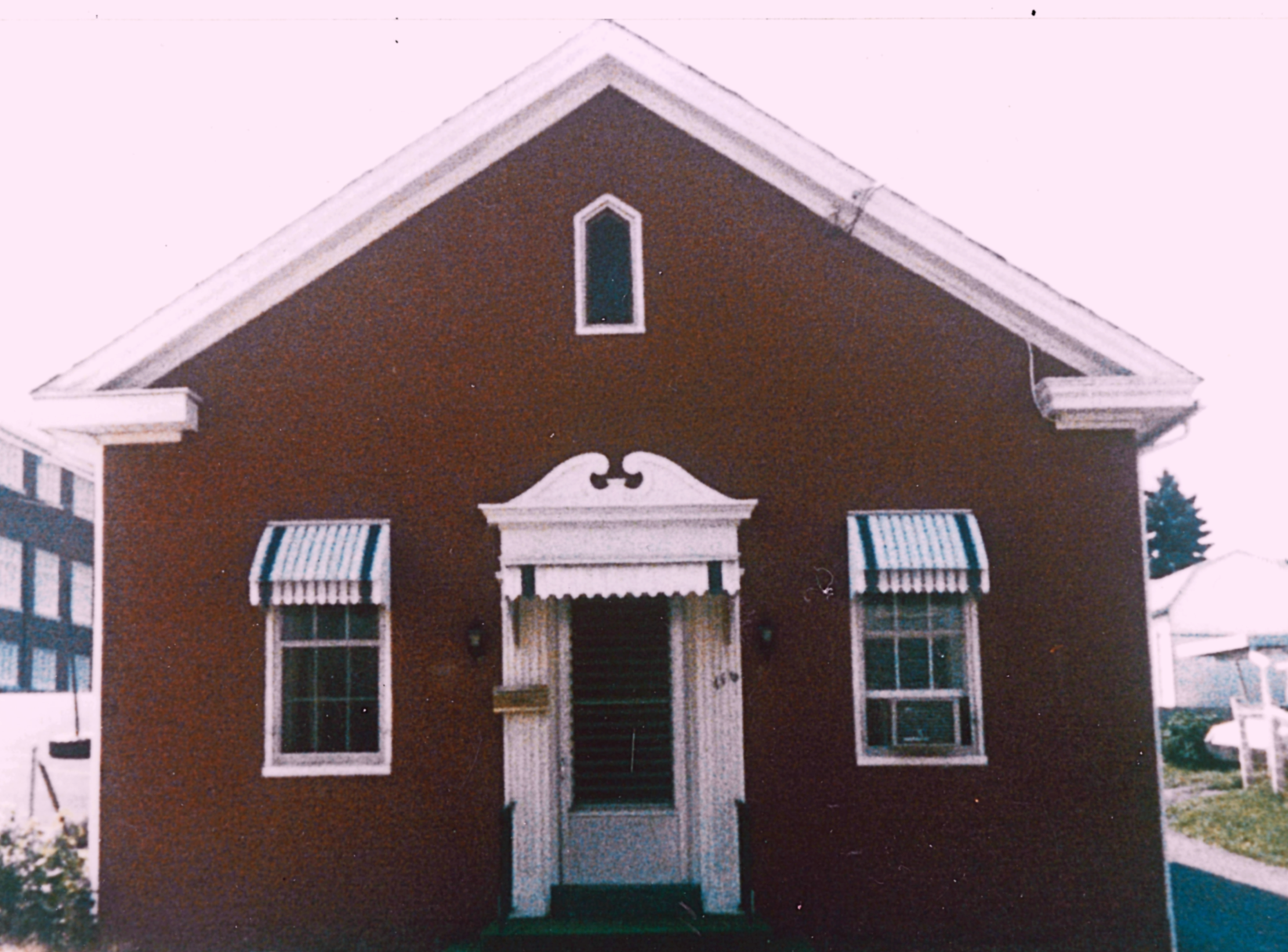 A red house with white trim and a gabled roof, featuring two windows with striped awnings and an entrance door with a decorative white pediment, surrounded by neighboring houses and a lawn.