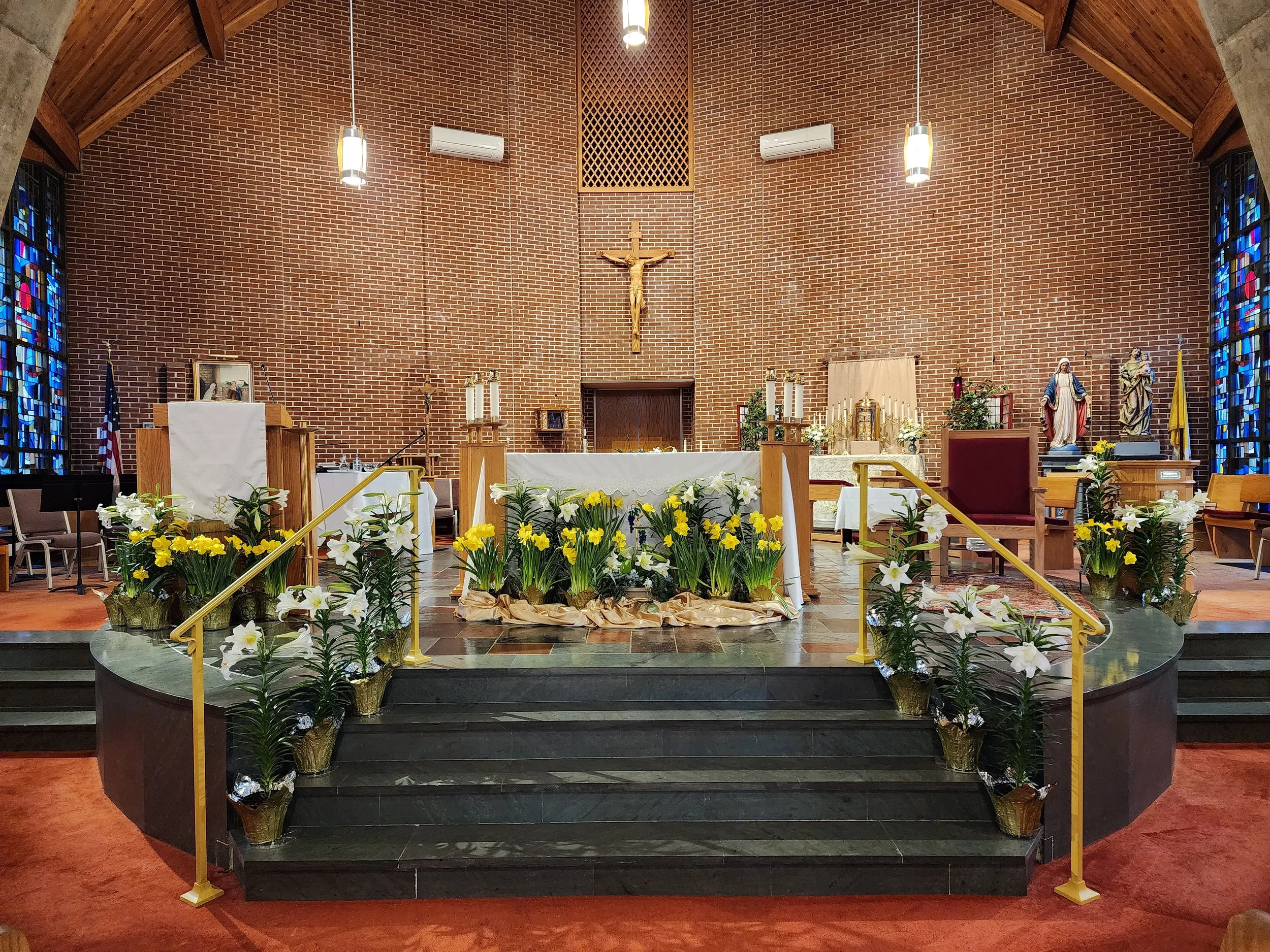 Interior of a church altar decorated with yellow and white flowers, with steps leading up to it. There are statues, candles, and religious symbols, including a crucifix on the brick wall behind the altar.
