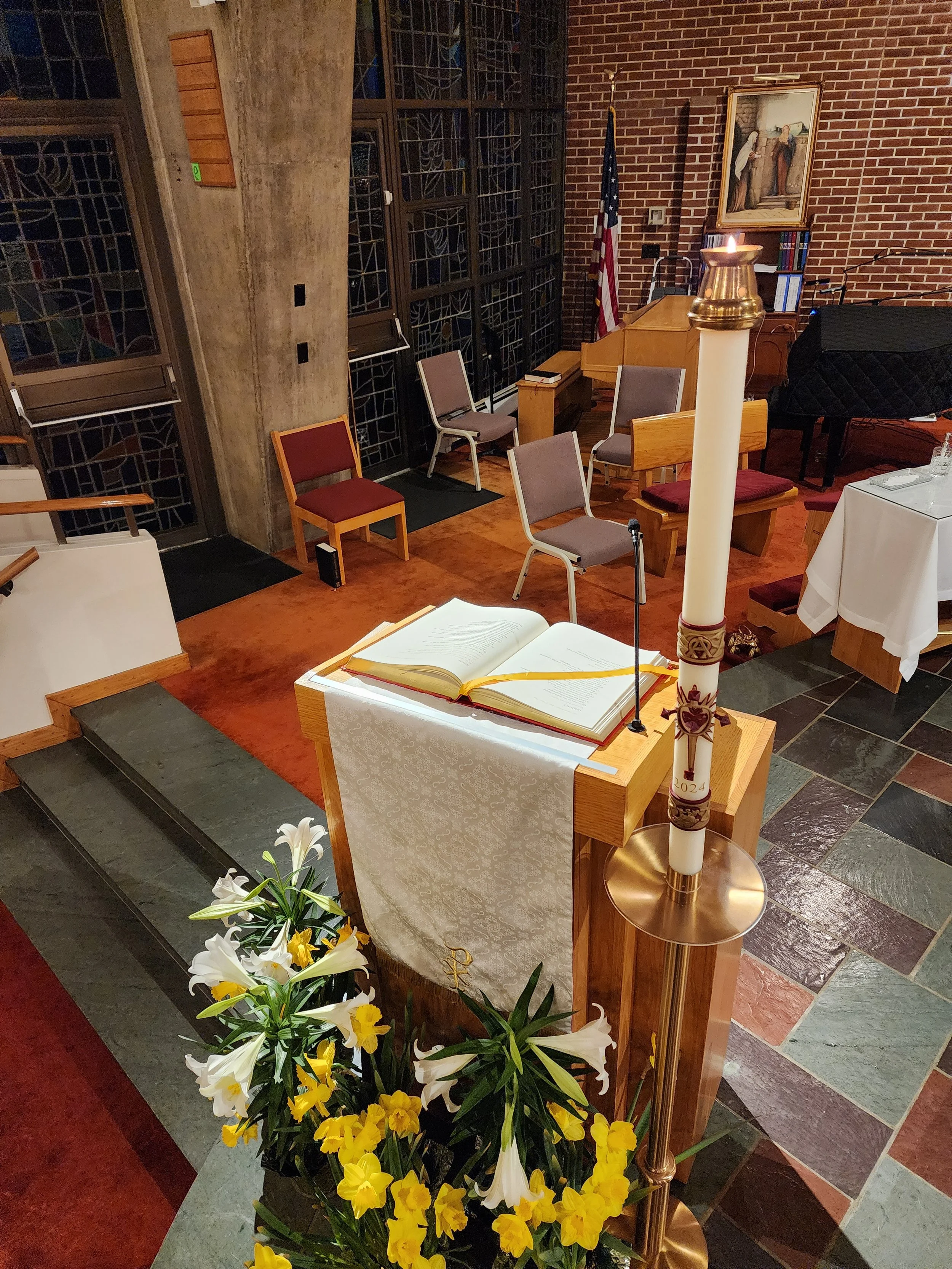 Interior of a church altar with a lit Paschal candle, an open Bible, and flowers, with chairs and stained glass windows in the background.