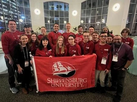 Group of twenty people holding a Shippensburg University banner, posing indoors with large windows behind them.