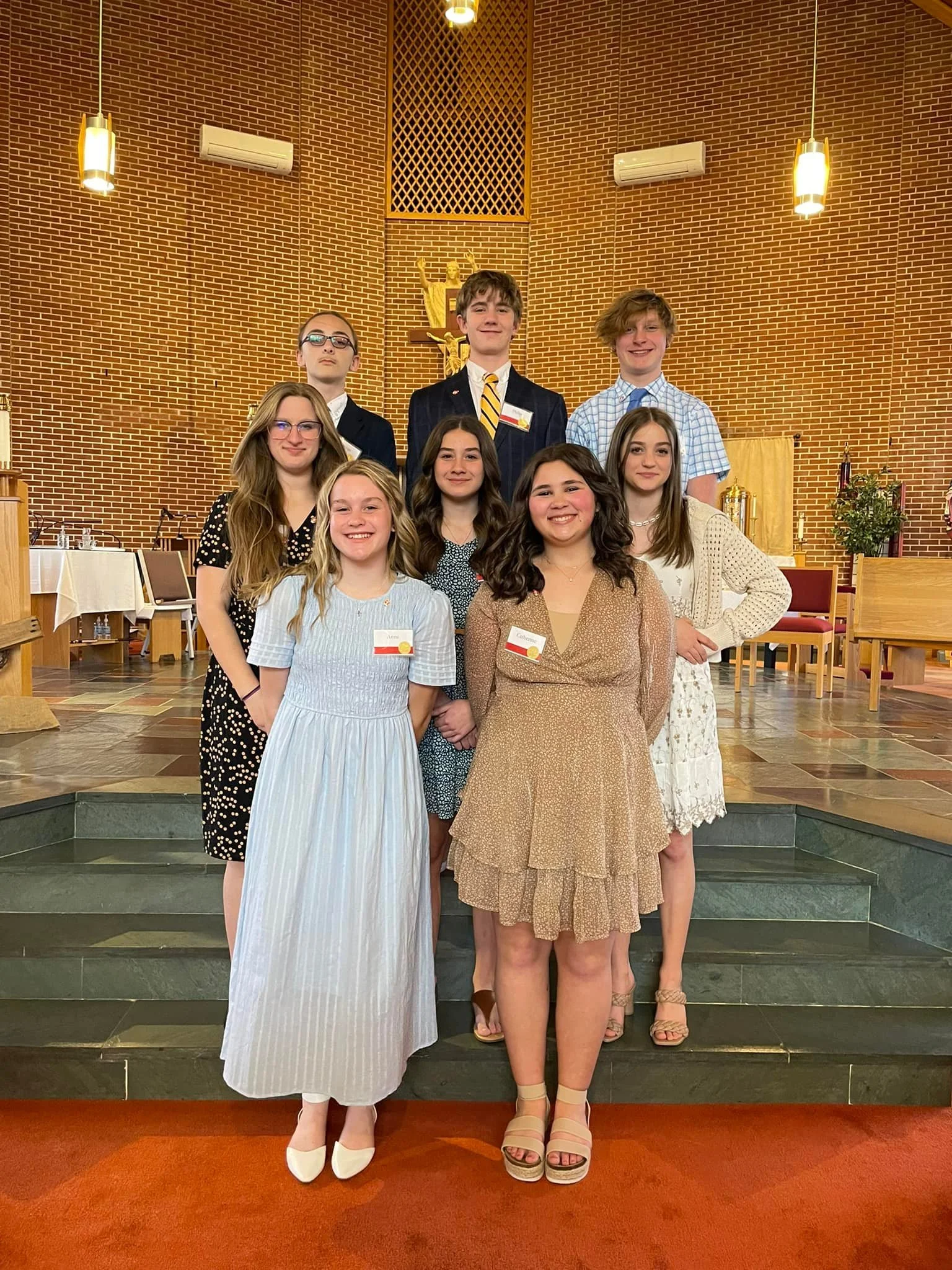 Group of nine young people standing together on steps inside a church, dressed in semi-formal attire, smiling for a photo.
