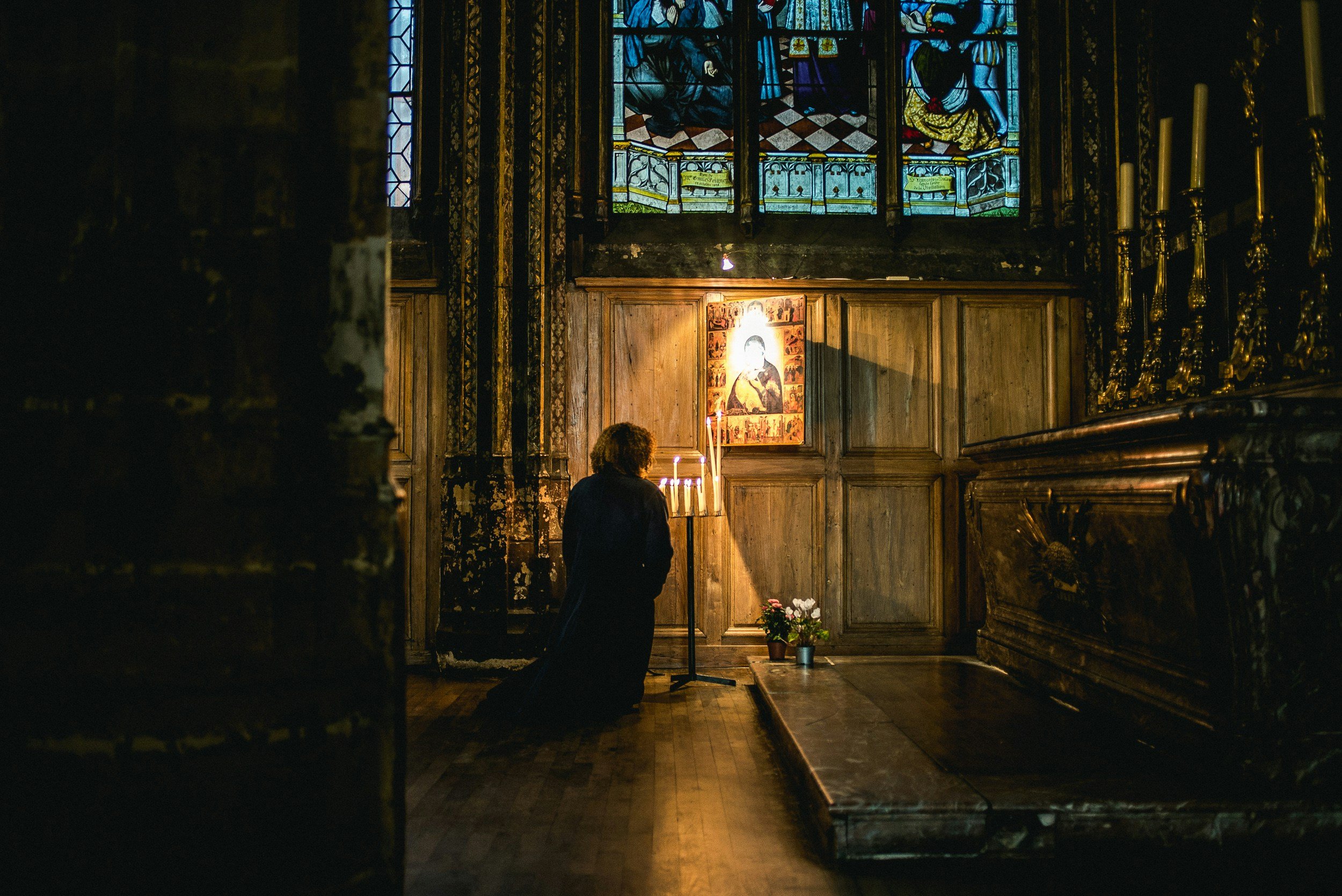 A woman kneeling in prayer inside a church, illuminated by candlelight. She is facing an icon and flowers, with stained glass windows above.