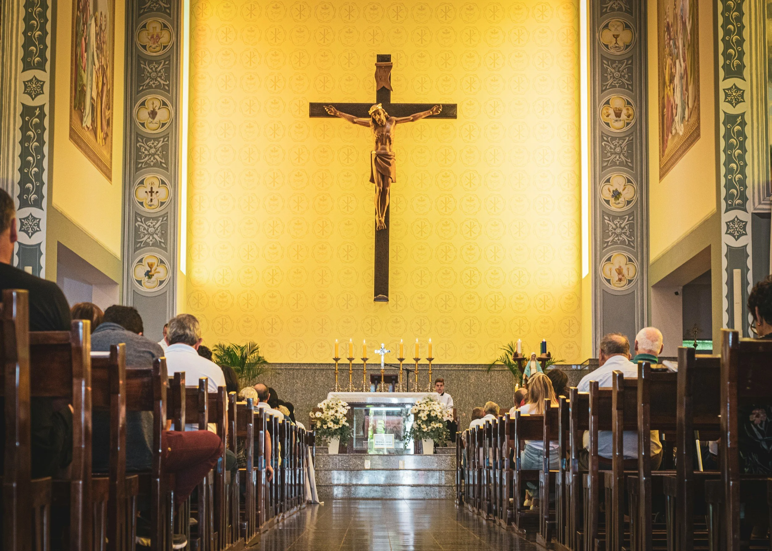 Inside a church during a service with people seated in pews facing an altar. A large crucifix hangs on a yellow wall behind the altar, which is decorated with flowers and candles.