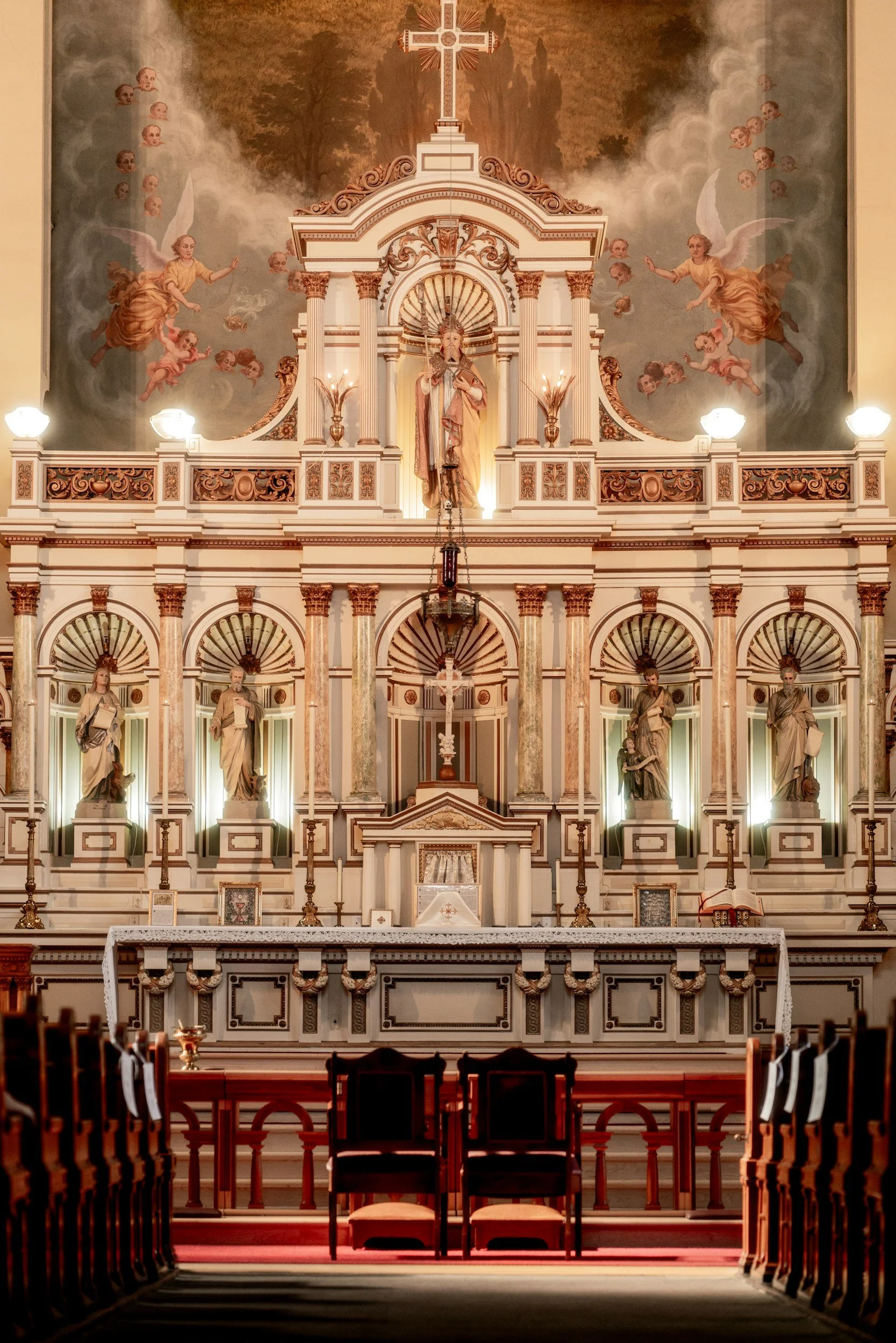 Interior of a church altar with statues, a crucifix, and religious artwork, featuring wood and marble details and soft lighting.