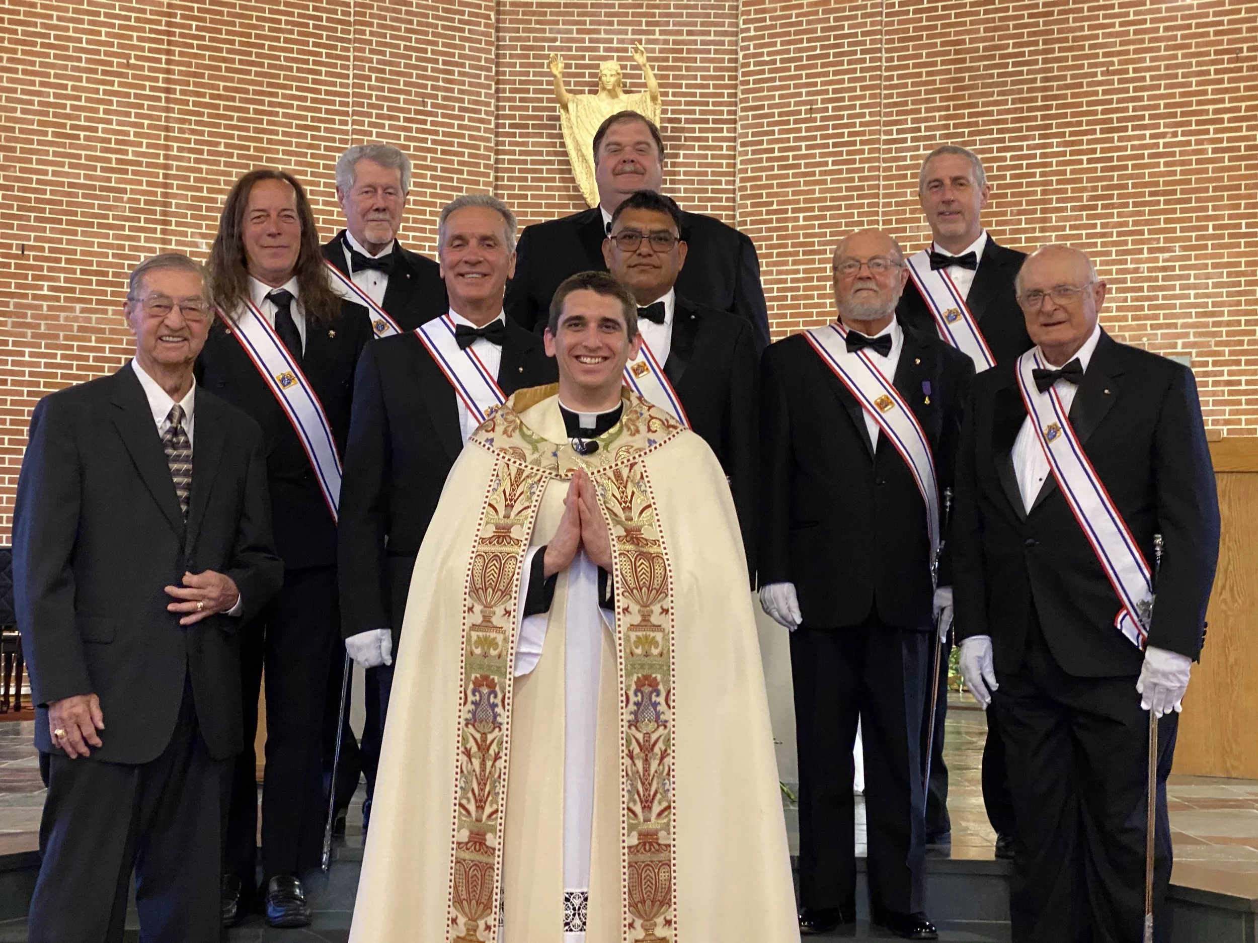 Group of men in tuxedos, a priest in ornate robes, and a woman in a clerical collar standing on a stage in a church with a brick wall and religious statue behind them.