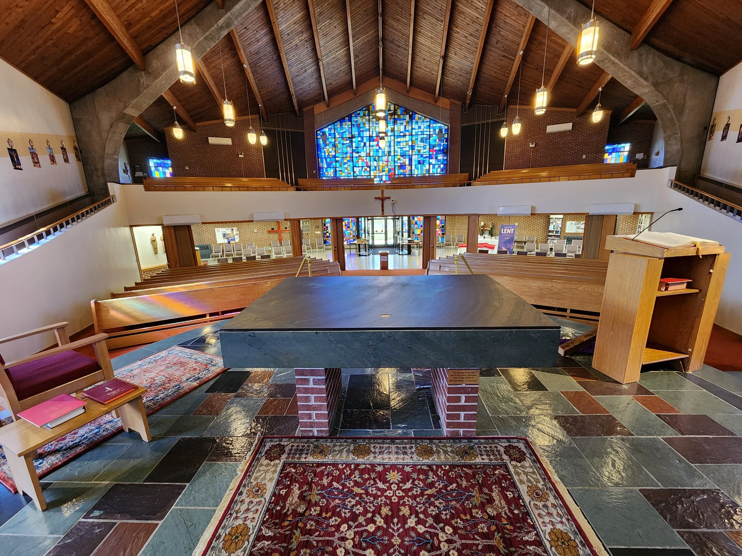 Interior view of a church altar with wooden benches, a pulpit on the right, colorful stained glass windows, hanging pendant lights, and a rug on the tiled floor.