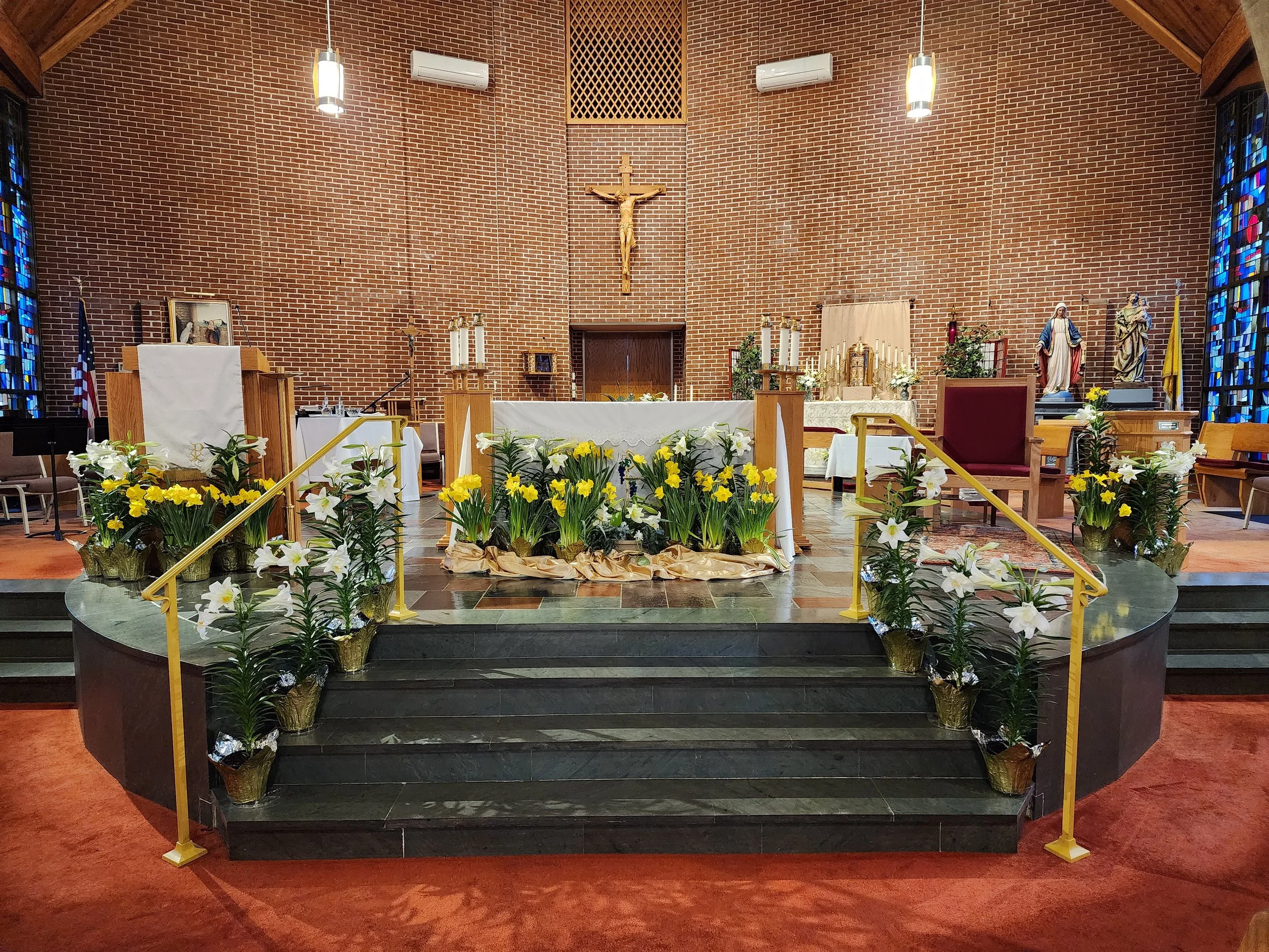 Interior of a church altar decorated with white and yellow flowers, with crucifix and statues of saints, brass votive candles, and stained glass windows.