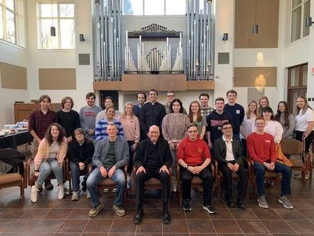 Group photo of 19 people, including men and women of various ages, standing and sitting inside a church with a pipe organ in the background.
