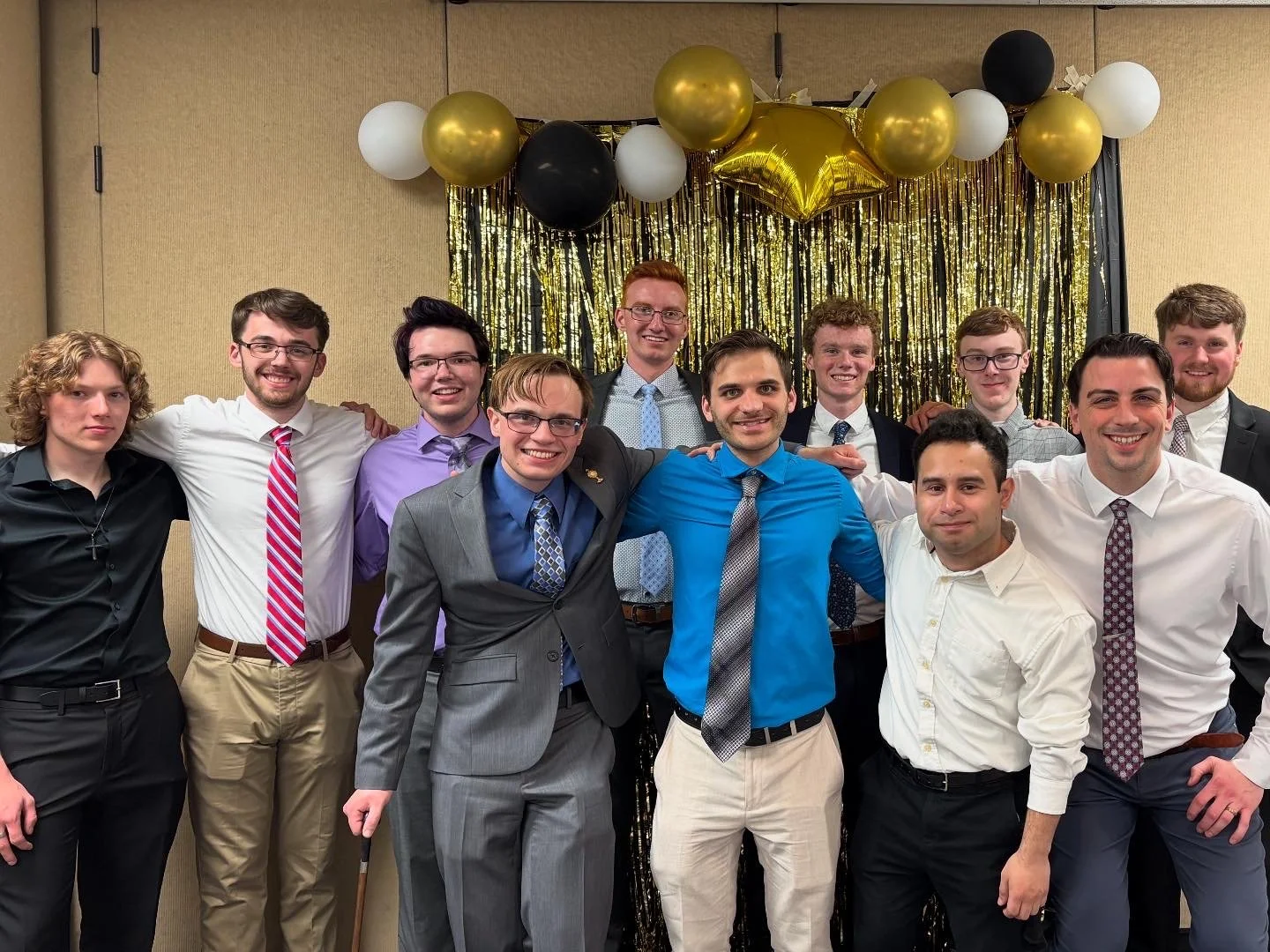 A group of twelve young men in formal attire posing for a photo at a celebration with gold, black, and white balloons and a gold fringe backdrop.