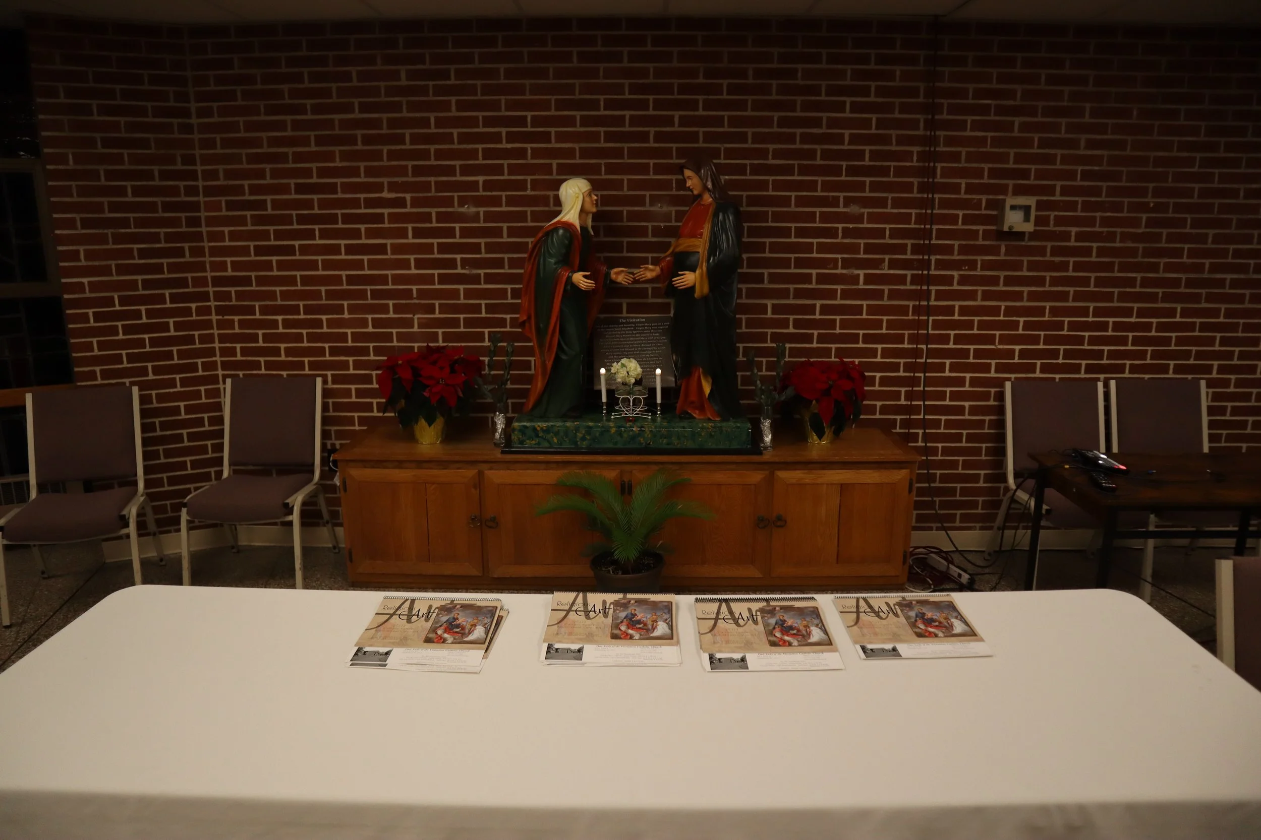 Religious altar with statues of Mary and Jesus Christ, poinsettia plants, magazines on tables, and chairs against a brick wall.