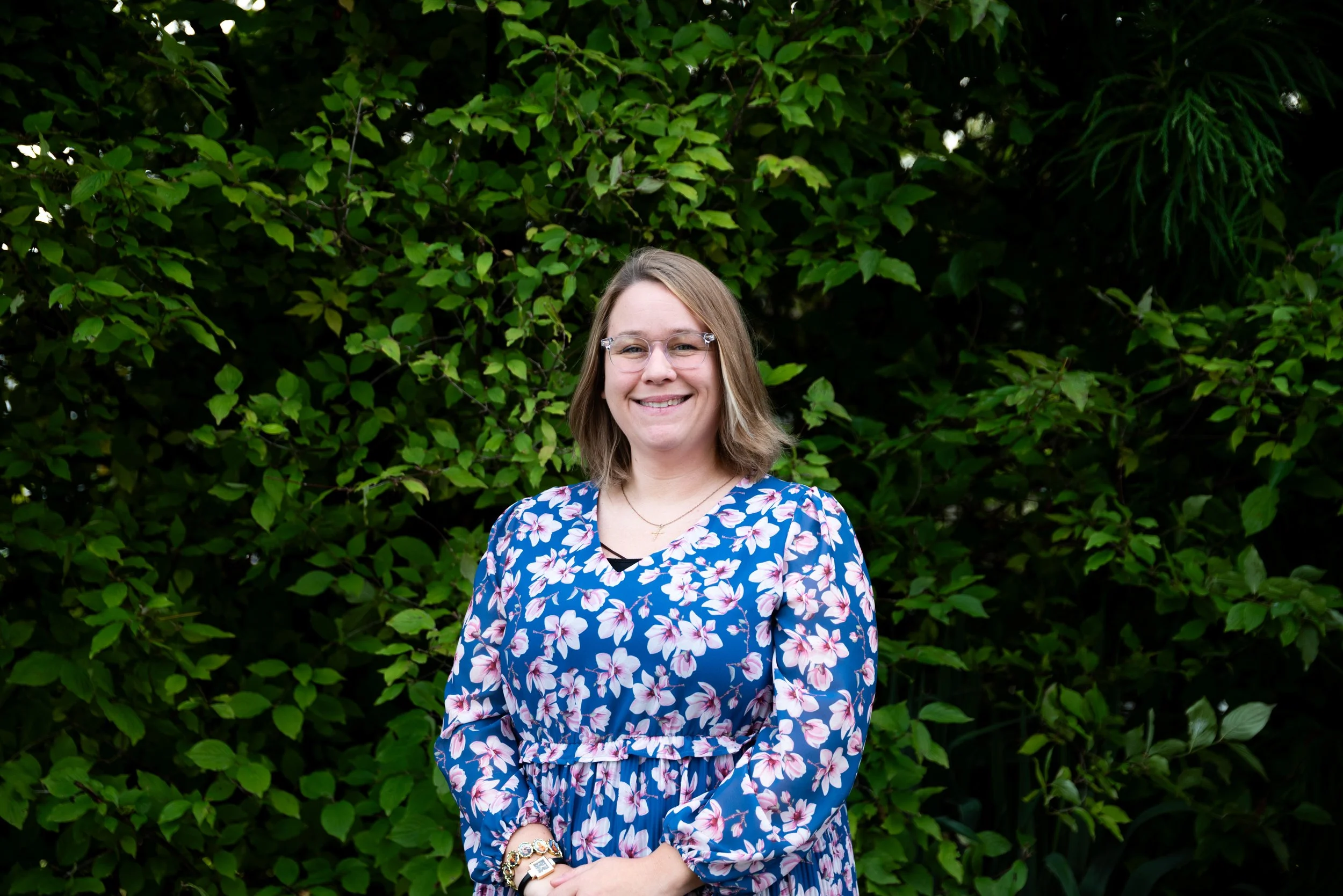 A woman in a blue floral dress smiling while standing outdoors in front of green shrubbery.