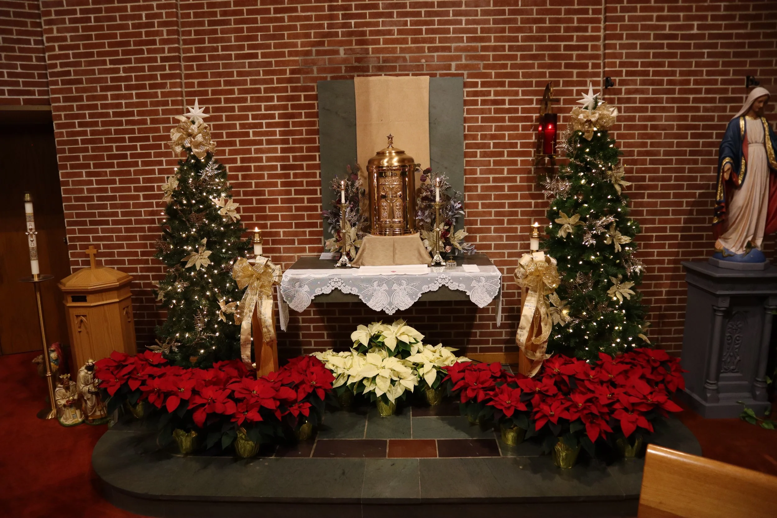 Christmas altar with two decorated Christmas trees, poinsettias, and religious statues in a brick church interior.