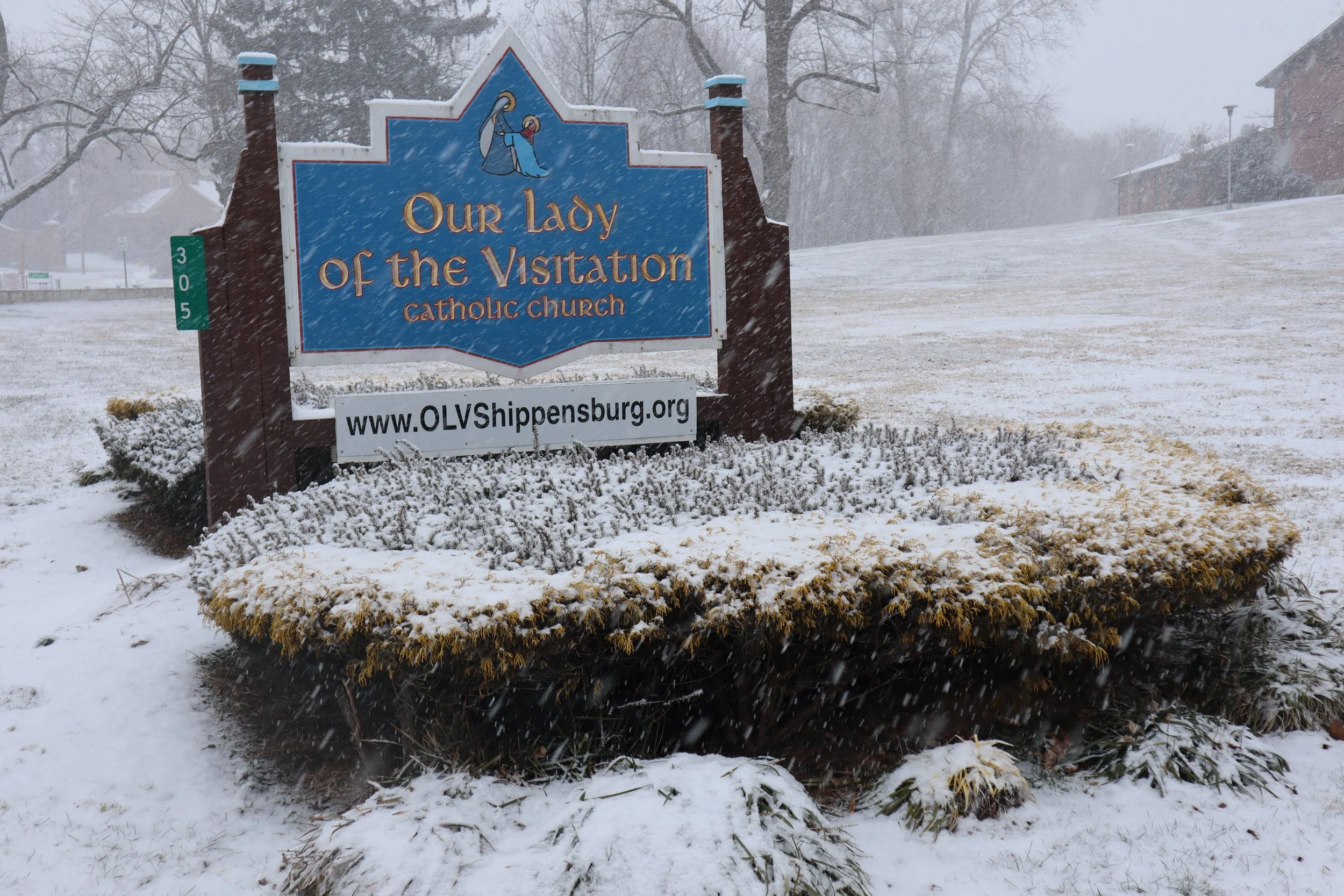 Snow-covered sign for Our Lady of the Visitation Catholic Church and website www.OLVShippensburg.org with snow falling in the background.