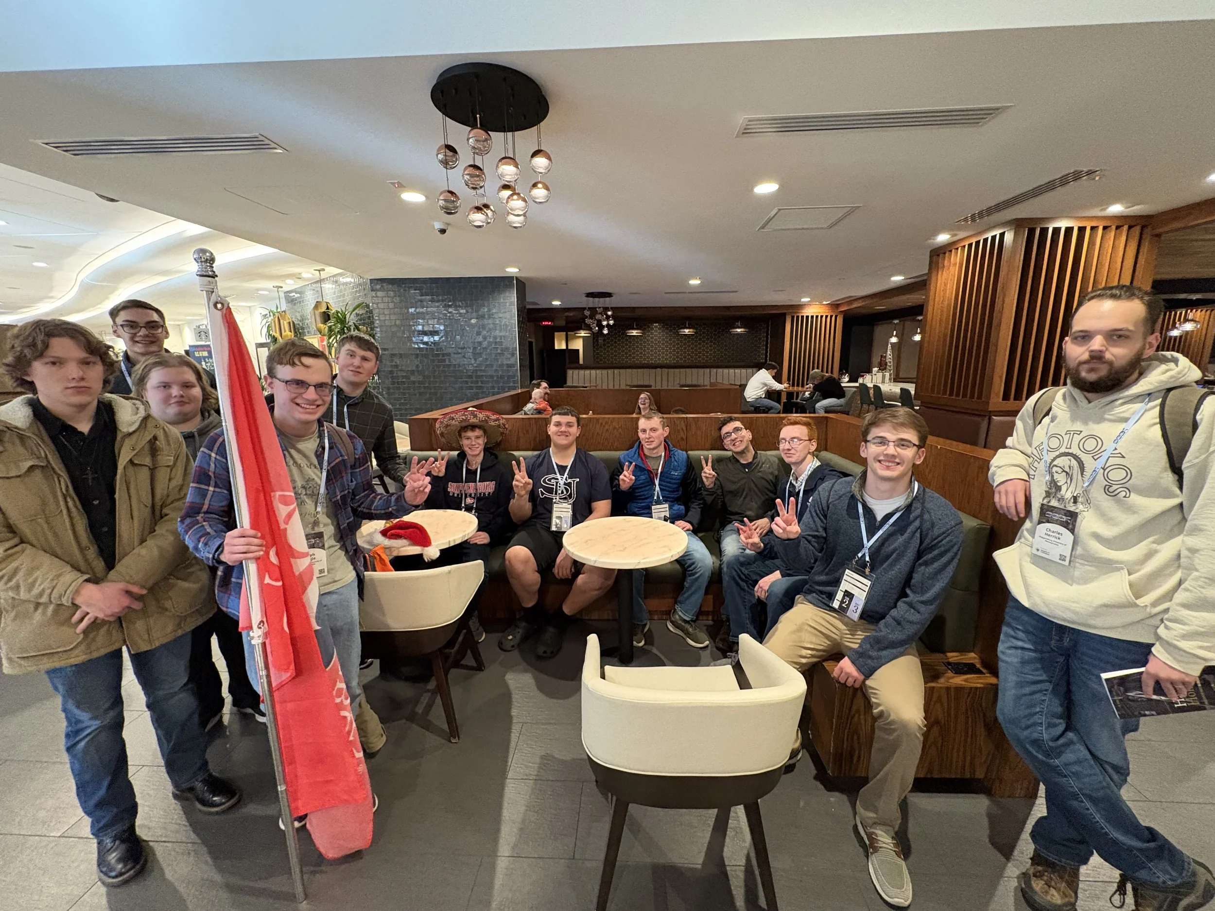 Group of young people, mostly smiling and making peace signs, gathered around a table in a restaurant or cafe, with some seated and others standing, in an indoor setting.