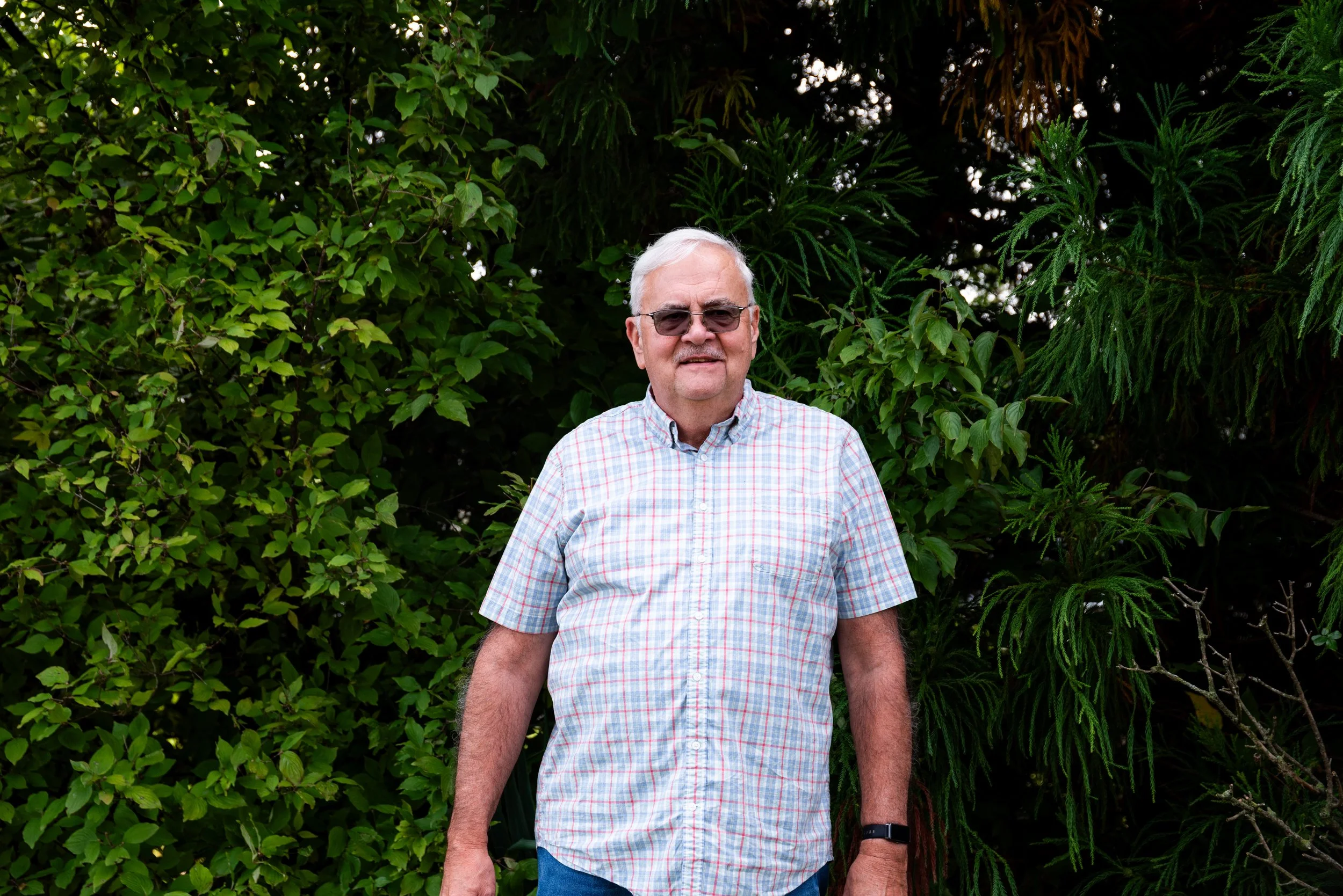 An elderly man with white hair and glasses standing outdoors in front of green foliage, wearing a short-sleeved plaid shirt and a watch.